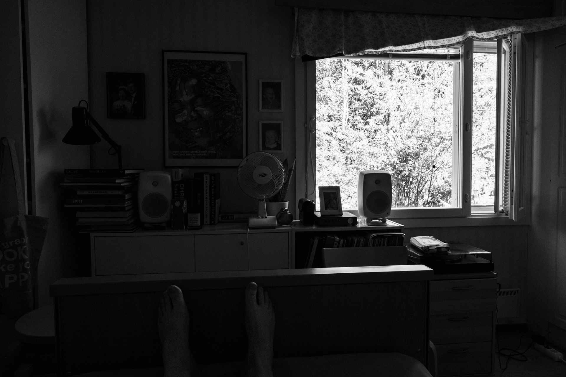 A black-and-white photograph of a view from a bedroom window to outside, with trees and vegetation filling the window view. The small side window is open. A sideboard has a fan on top of it.