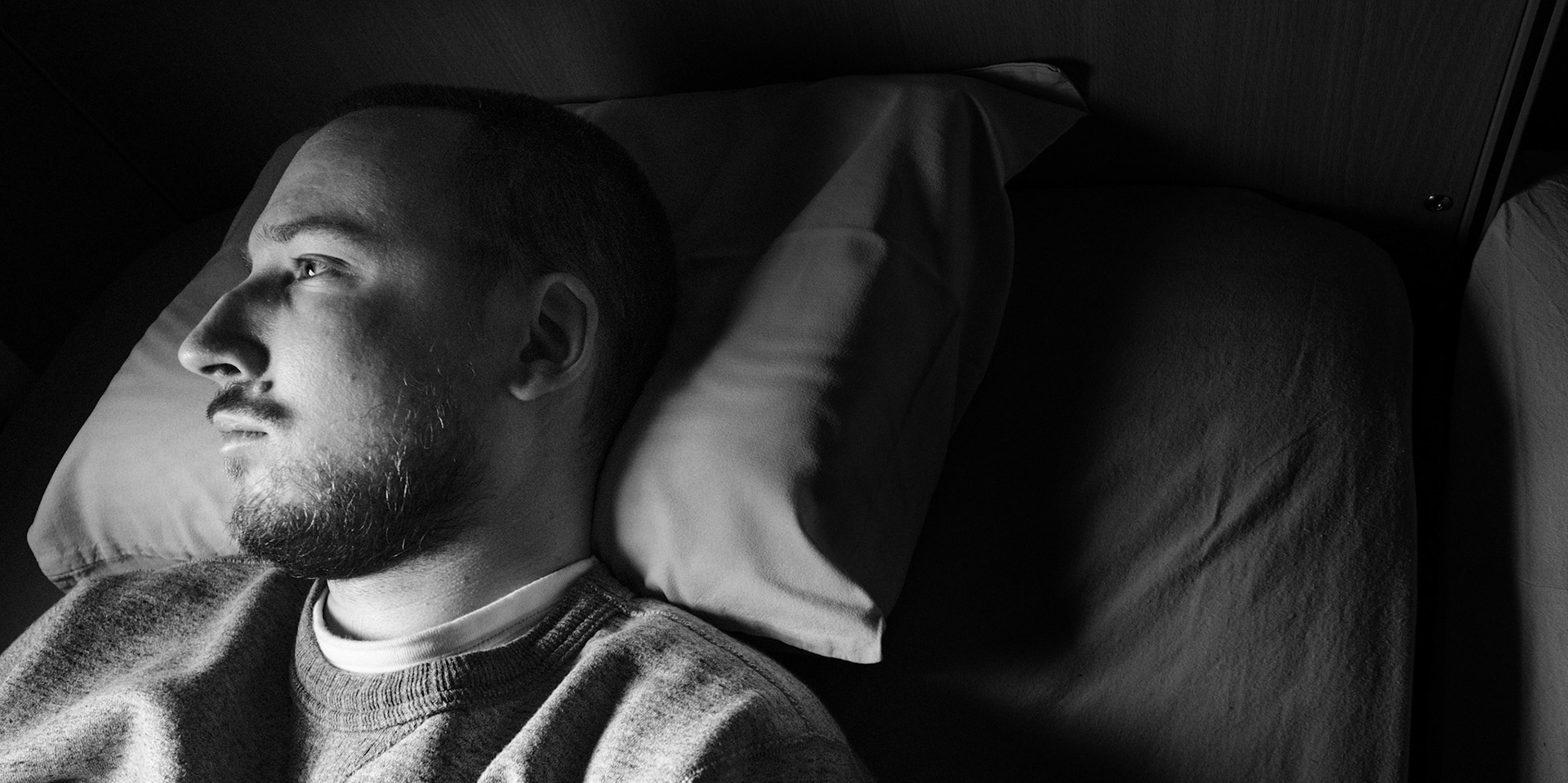 A high contrast black and white close-up of a man, Niko, resting his head on a pillow, gazing outside the frame. High-contrast lighting highlights his facial features, including his Roman nose, while the rest of the environment remains dark.