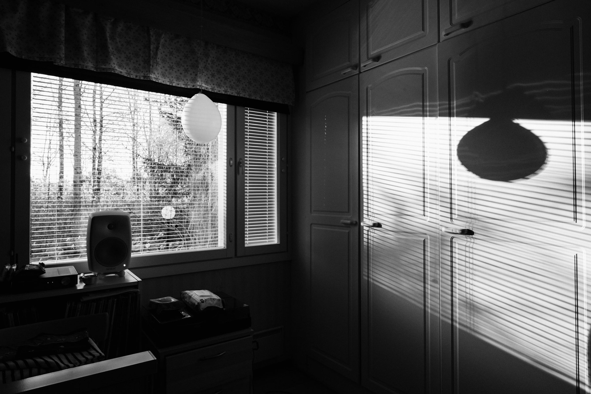 A black-and-white photo of a window view revealing a springy landscape. Inside the room, there is a balloon hanging in front of the window, which casts a long shadow onto a closet. There is also a turntable and a speaker, and various personal belongings on top of a sideboard.