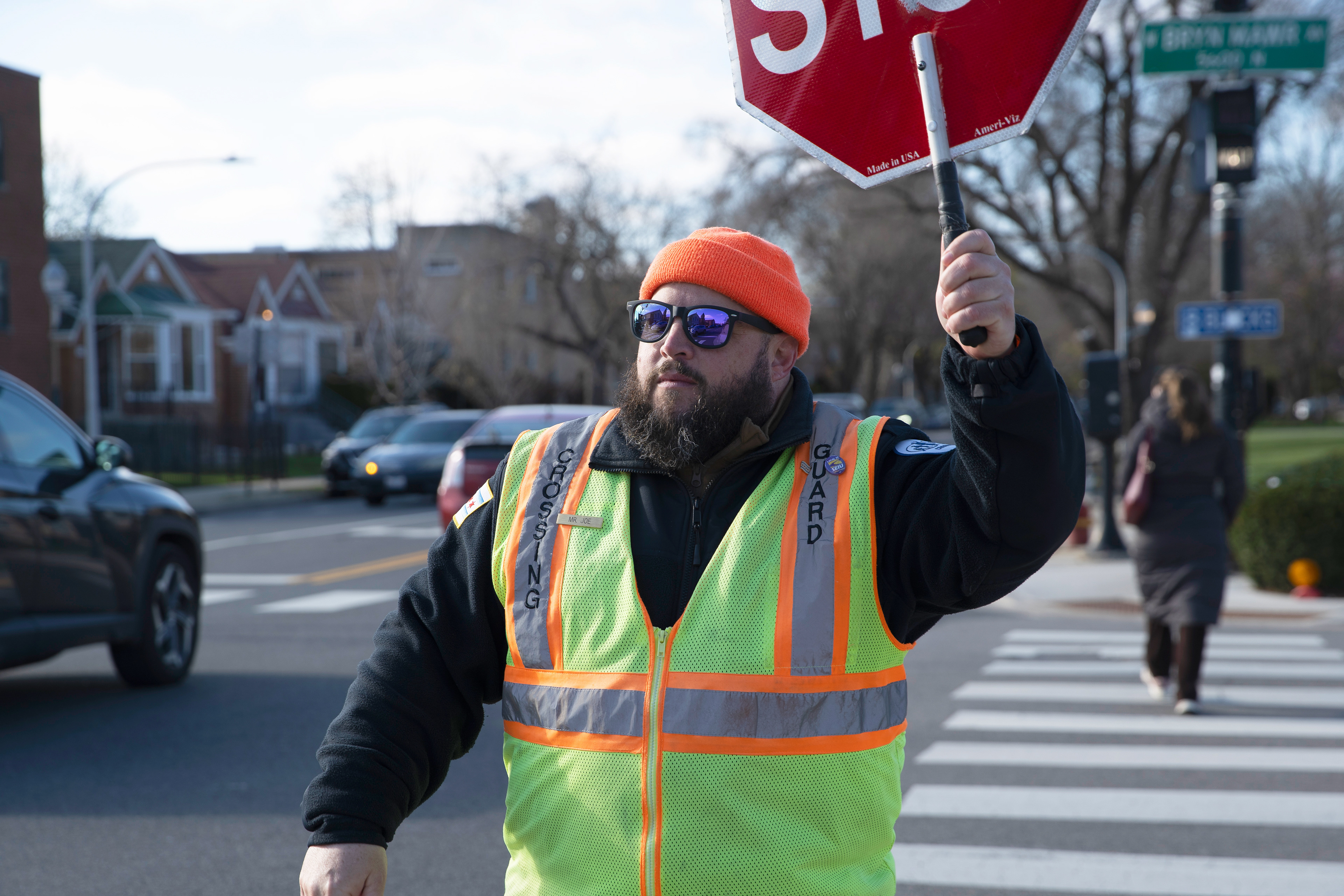 Chicago Public Schools crossing guard Joe Sass marches across a cross walk holding a large stop sign in Chicago on April 7, 2026. According to Sass, he helps roughly 105 pedestrians cross his intersection during his morning 35-minute shift.