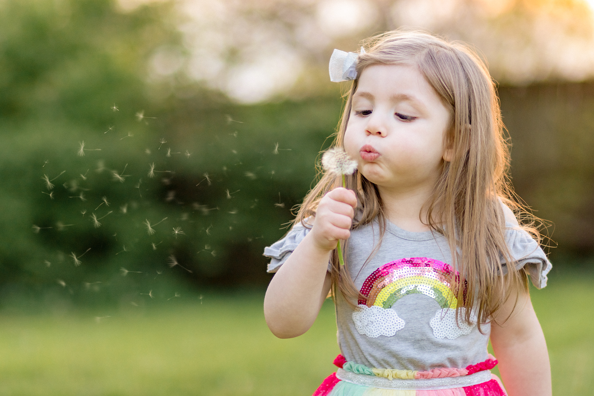Photo of a child in Cyntihana Kentucky wearing a rainbow dress blowing a dandelion