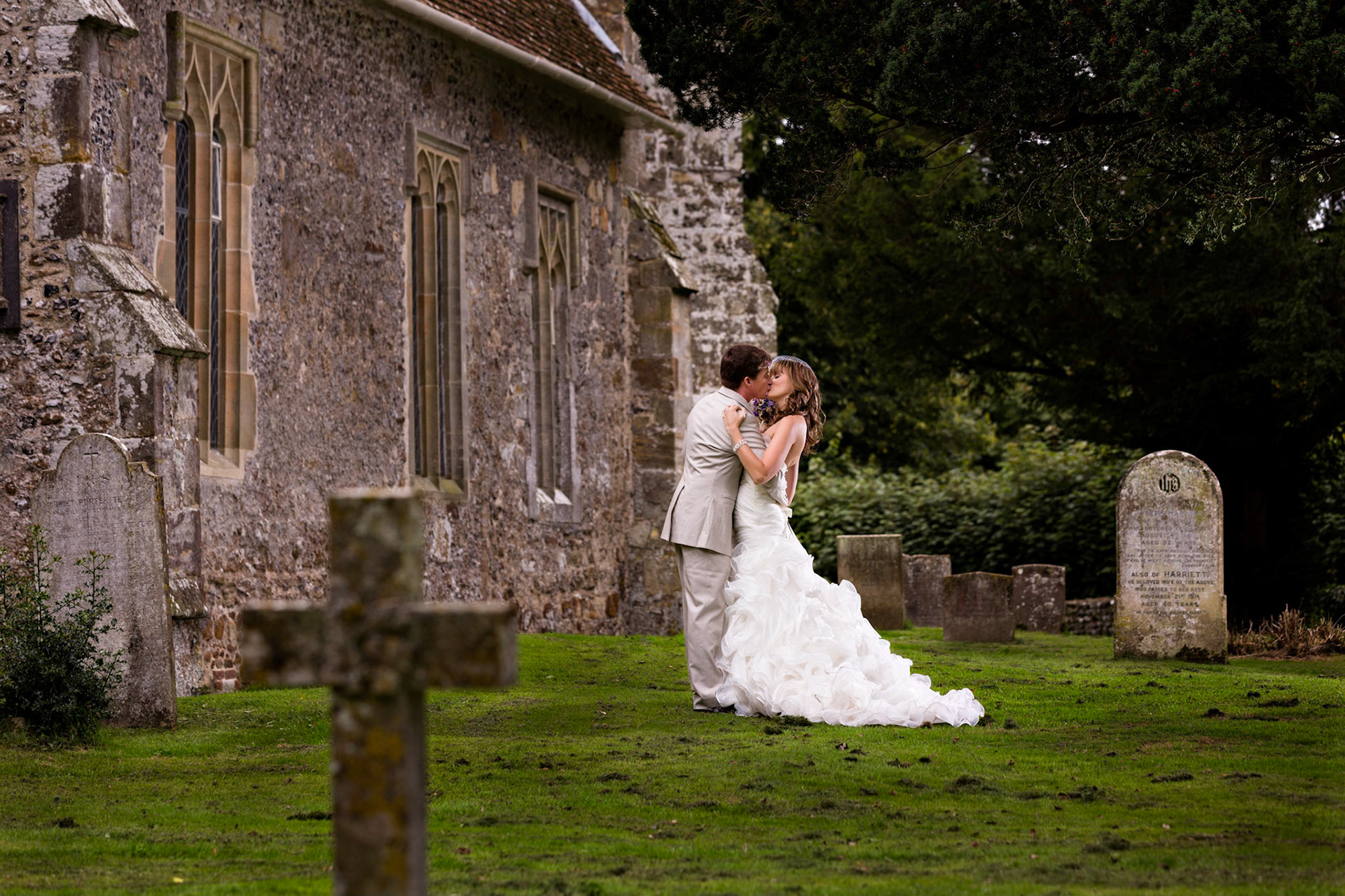 Tim and Samantha, the bride and groom, kiss outside the St John the Baptist Church in Ripe England after their destination wedding. This little church is so old that it is in the Dooms Day Book.