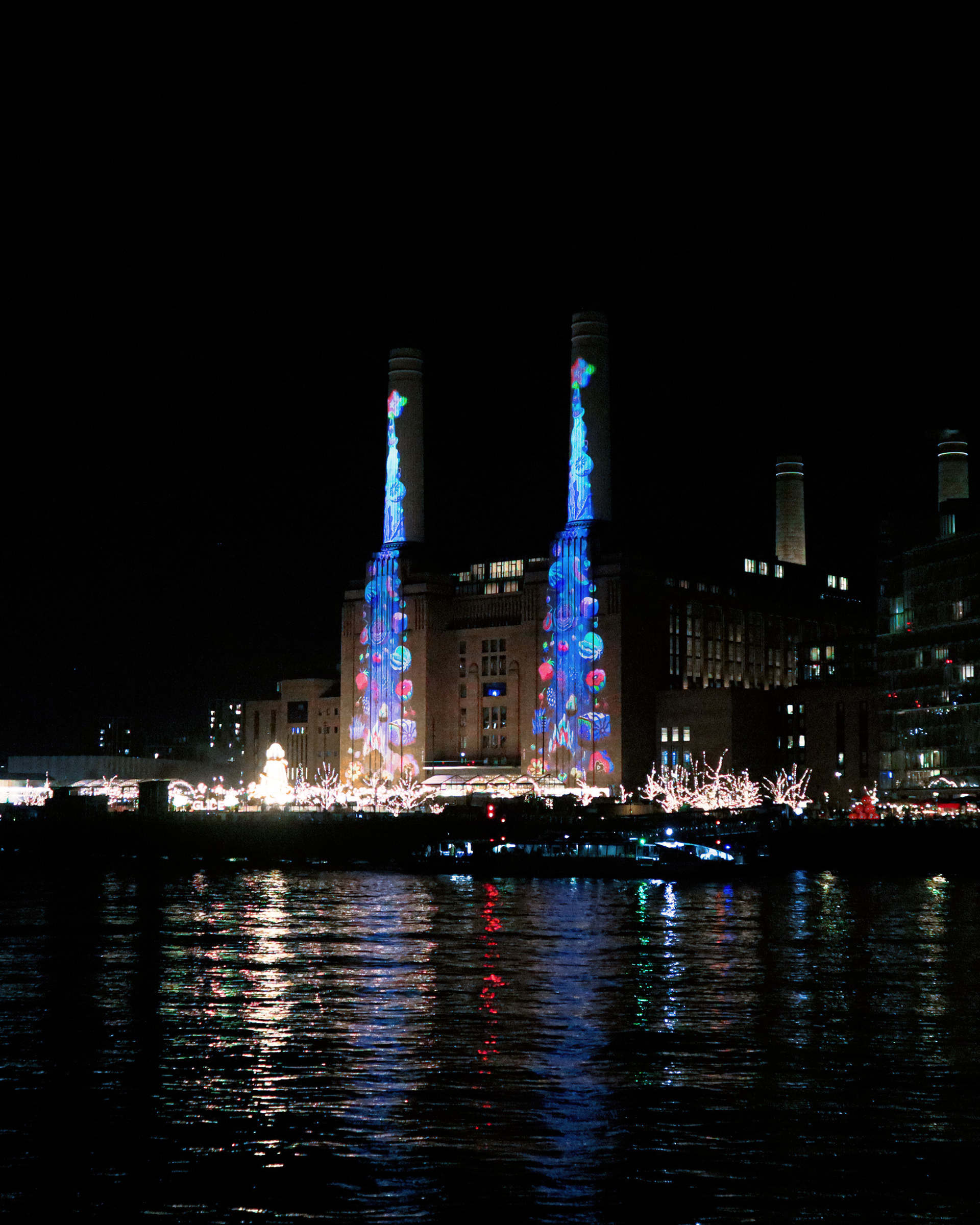 Large-scale public art projection by Sam Wilde illuminating the chimneys of Battersea Power Station in London, reflected across the River Thames at night.