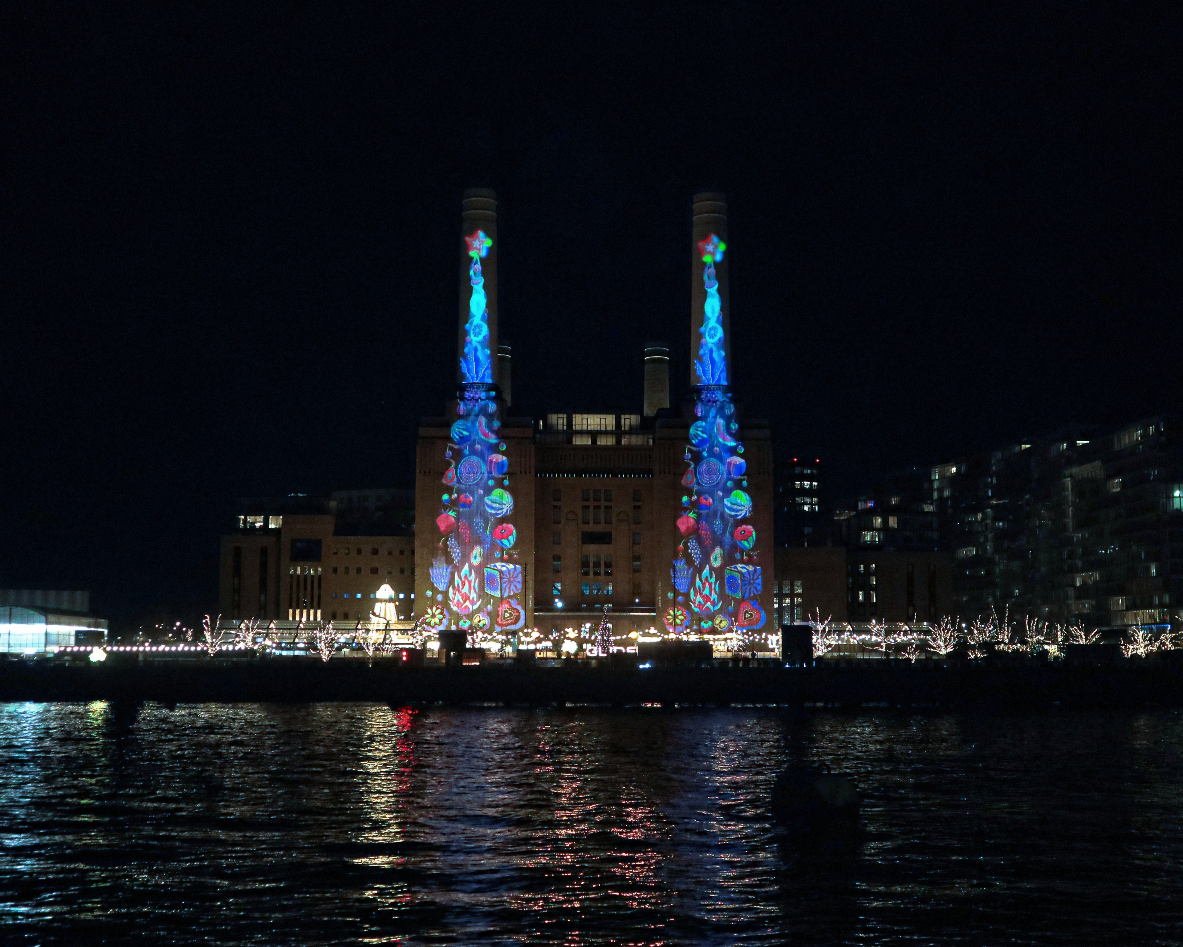 Night view of Battersea Power Station in London featuring a large-scale animated light artwork by Sam Wilde projected onto the twin chimneys, reflected across the River Thames.