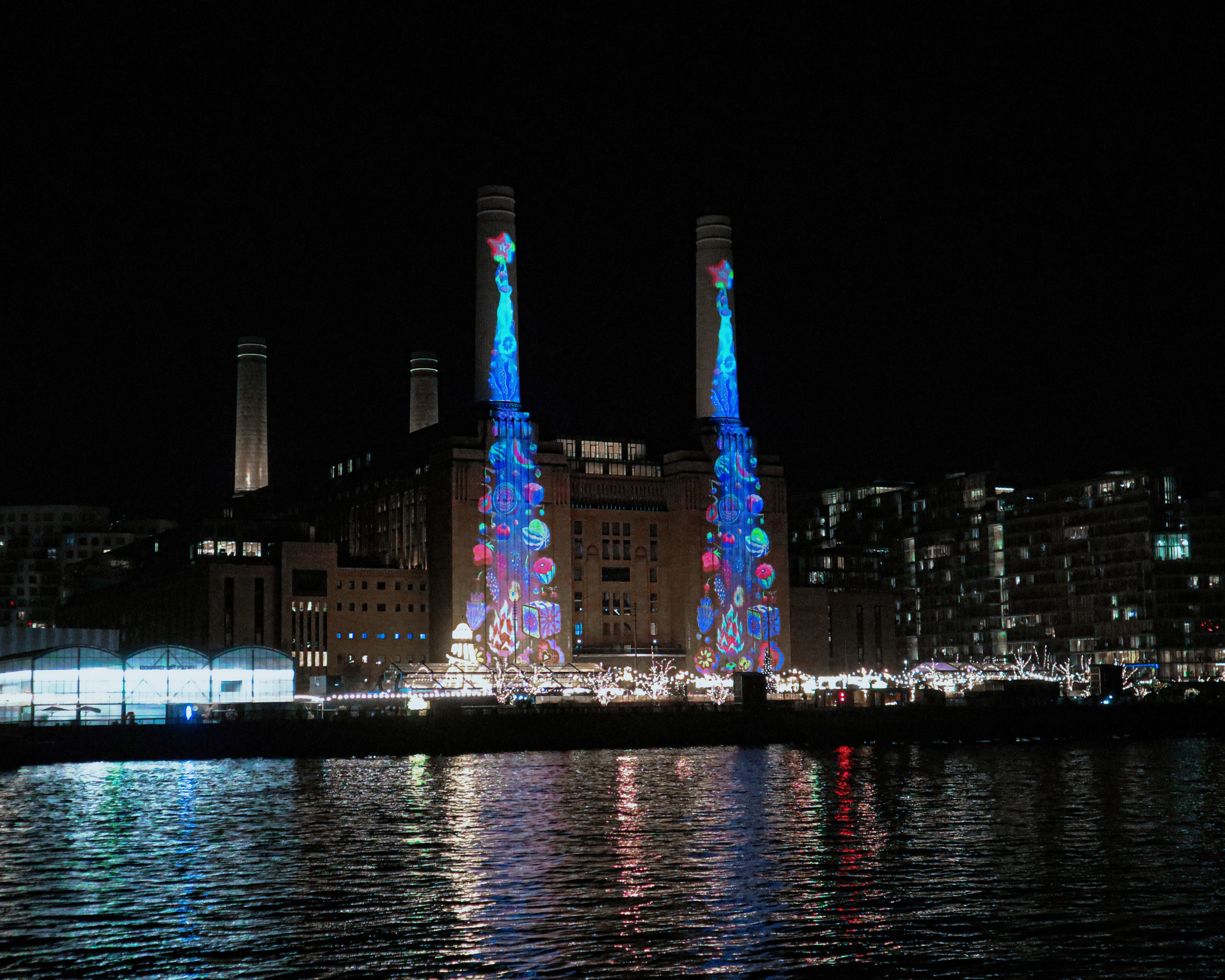Large-scale digital public art projection by Sam Wilde illuminating the chimneys of Battersea Power Station, London, set against the surrounding riverside architecture at night.