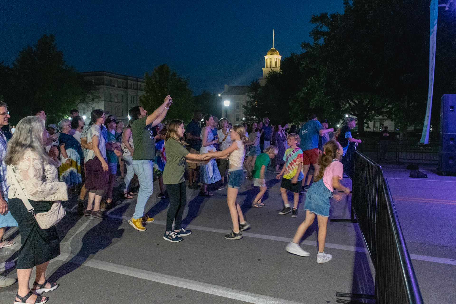 IOWA CITY, IOWA – JUNE 6, 2025: A multigenerational crowd fills the street in front of the Old Capitol as The Original Pinettes Brass Band performs nearby. Children dance, couples sway, and the community celebrates together under the summer night sky.