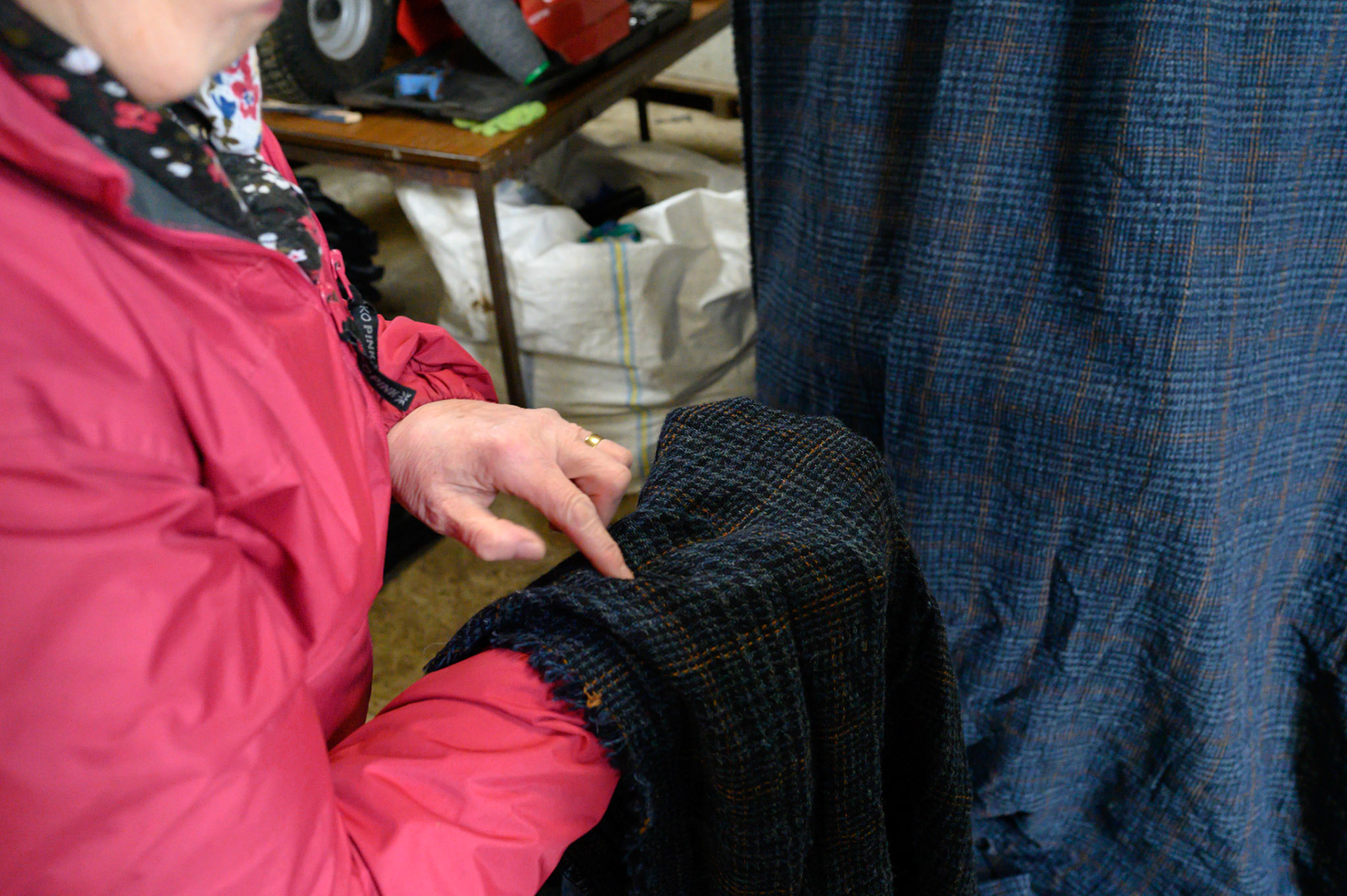 Anna Morrison points out the missing thread in the cloth she is working on.
Anna works as a darner for the Shawbost mills. She checks the lengths of Harris tweed for mistakes and loose threads. A lose thread sounds easier, as it must be only tied off and into the cloth.
This 70m length of Harris tweed is missing yellow thread from its pattern. Anna must weave the missing thread along the whole 70m with a needle. It is long painstaking work that needs a good eye and a patient mind.