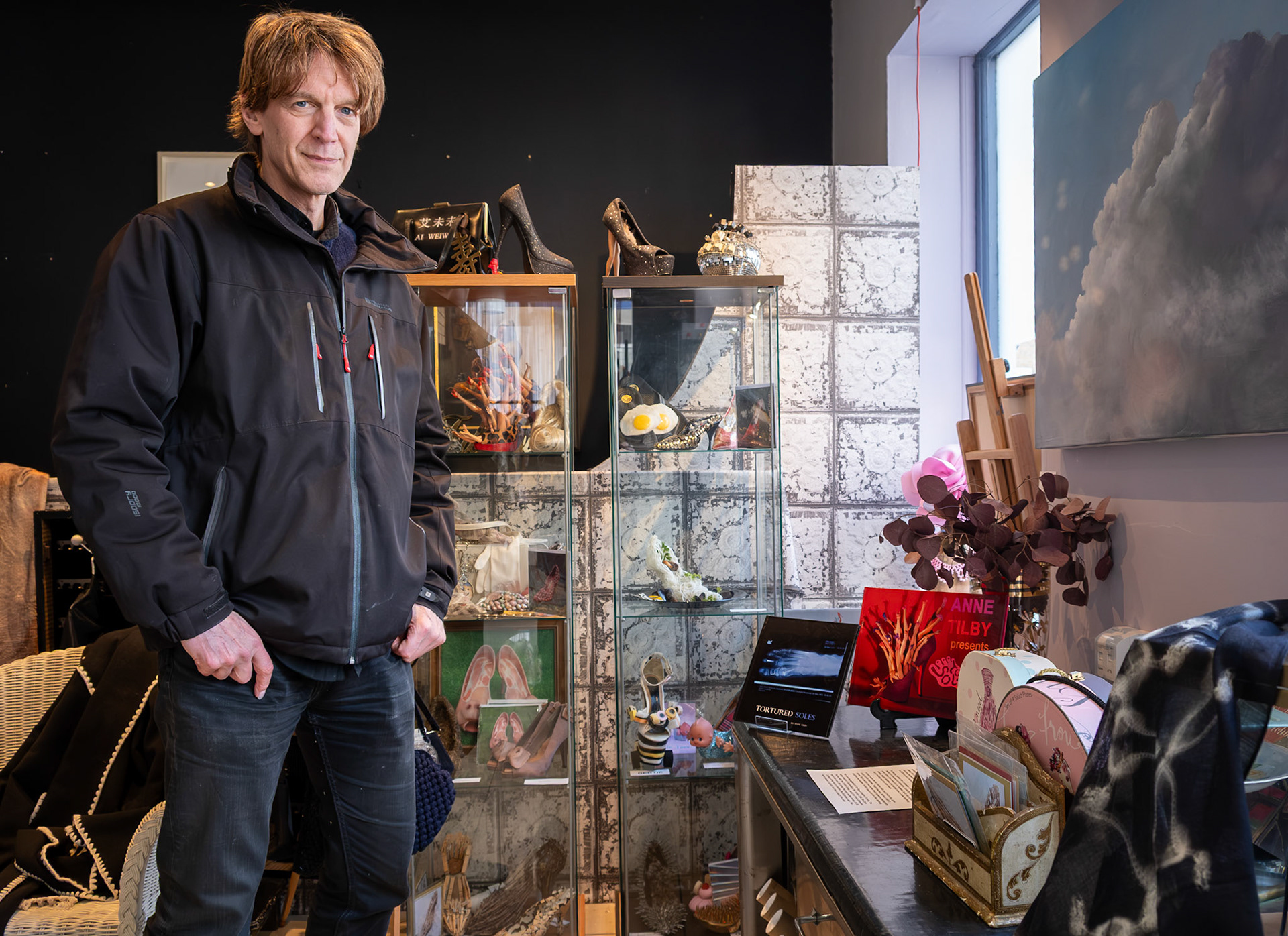 Graham Fink in his shop, 20 Cuckoos, 20 Market Street. 21st March, 2026. With their backgrounds in Advertising and Music, Graham Fink and his partner, Deirdre Allen, offer an eclectic mix of vintage clothing, furniture and objets d'art. The two books bottom right are by Anne Tilby and the display of humorous and unwearable footwear in the glass display cases are the original shoes featured in her book - Tortured Soles: the story of western foot binding. 