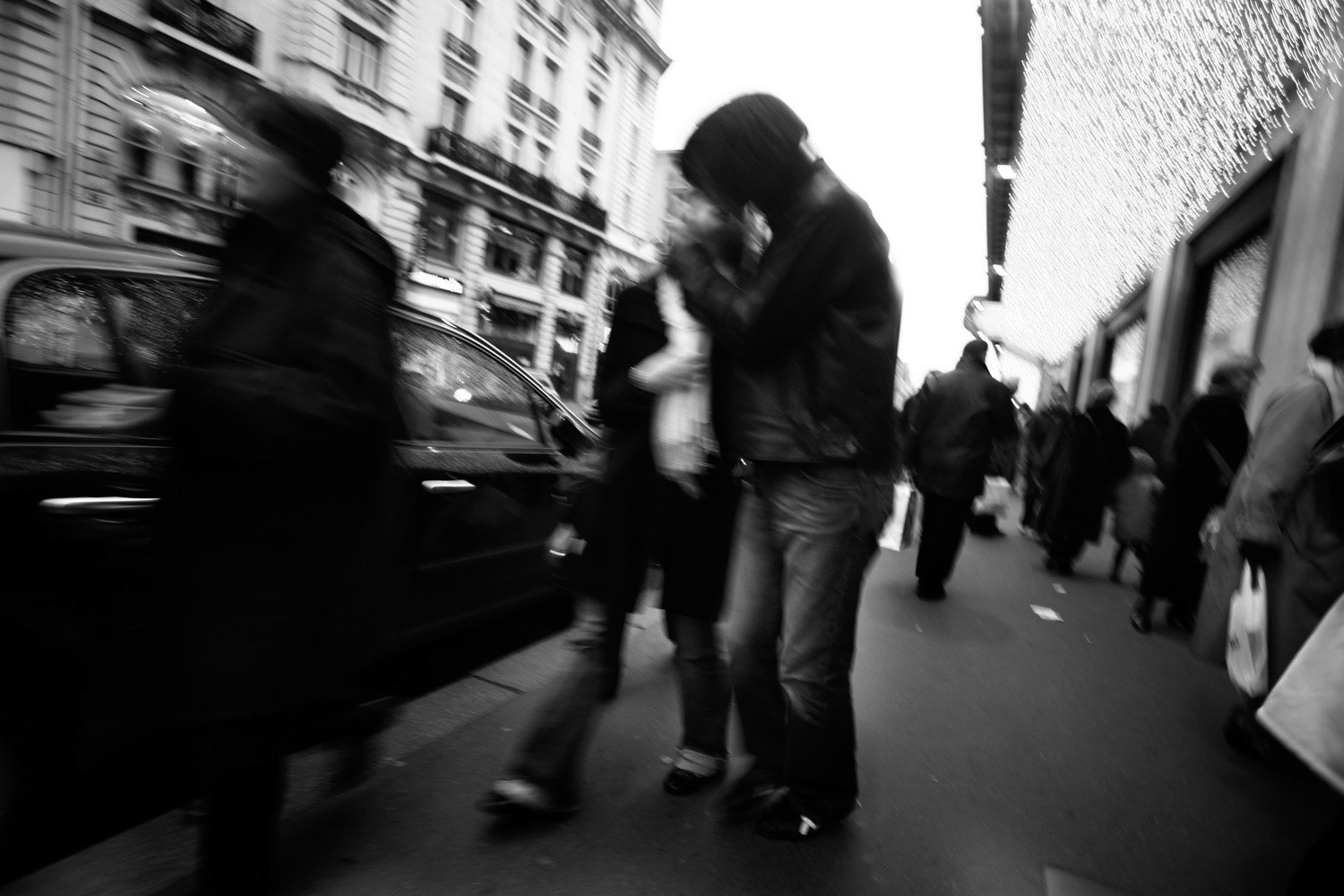 A Parisian couple kiss on the street at dusk as christmas lights line the street.