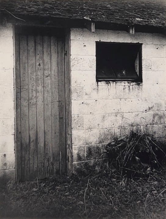Black and white photograph of the entrance to an abandoned house with the words "Keep out till payday" etched onto the slatted wooden door.