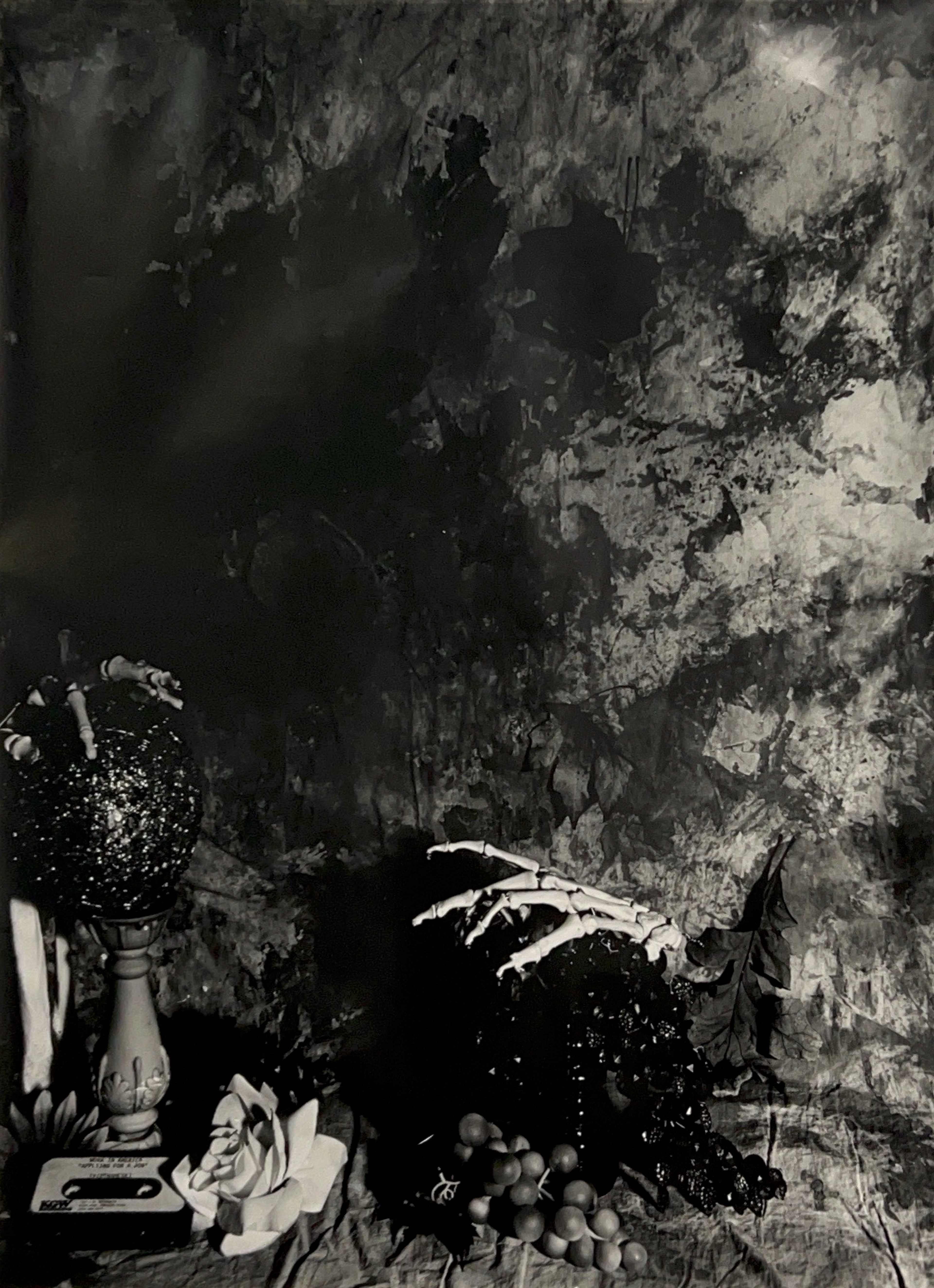 Black and white photograph of a still life scene with skeleton hands, grapes, a cassette tape, flowers, and a candle holder topped with a glittery ball. The backdrop is covered in smudges.