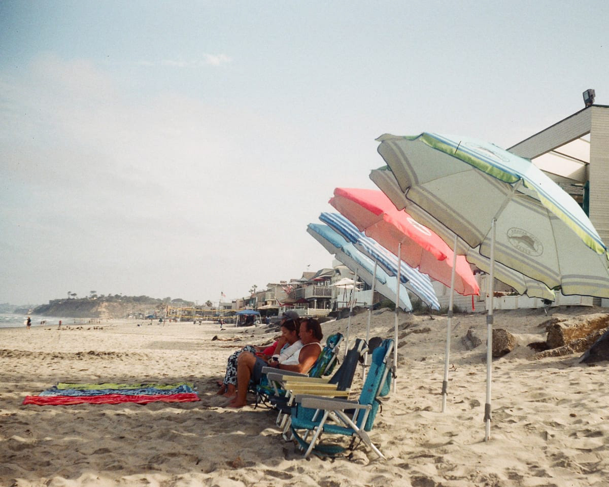Color photograph of people sitting in beach chairs under a row of colorful umbrellas in blue, red, and green colors.
