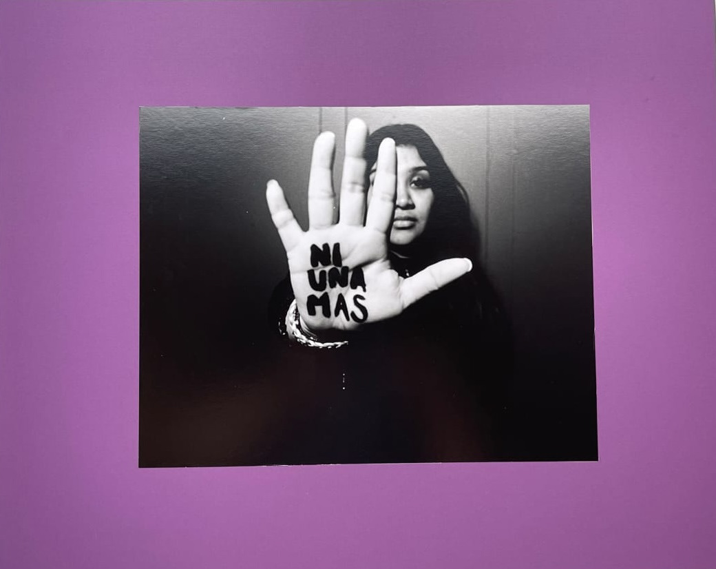 Black and white photo mounted on a purple board showing a woman holding her hand out to the camera in a "stop" motion with the words "Ni una mas" written on her hand in thick dark ink.