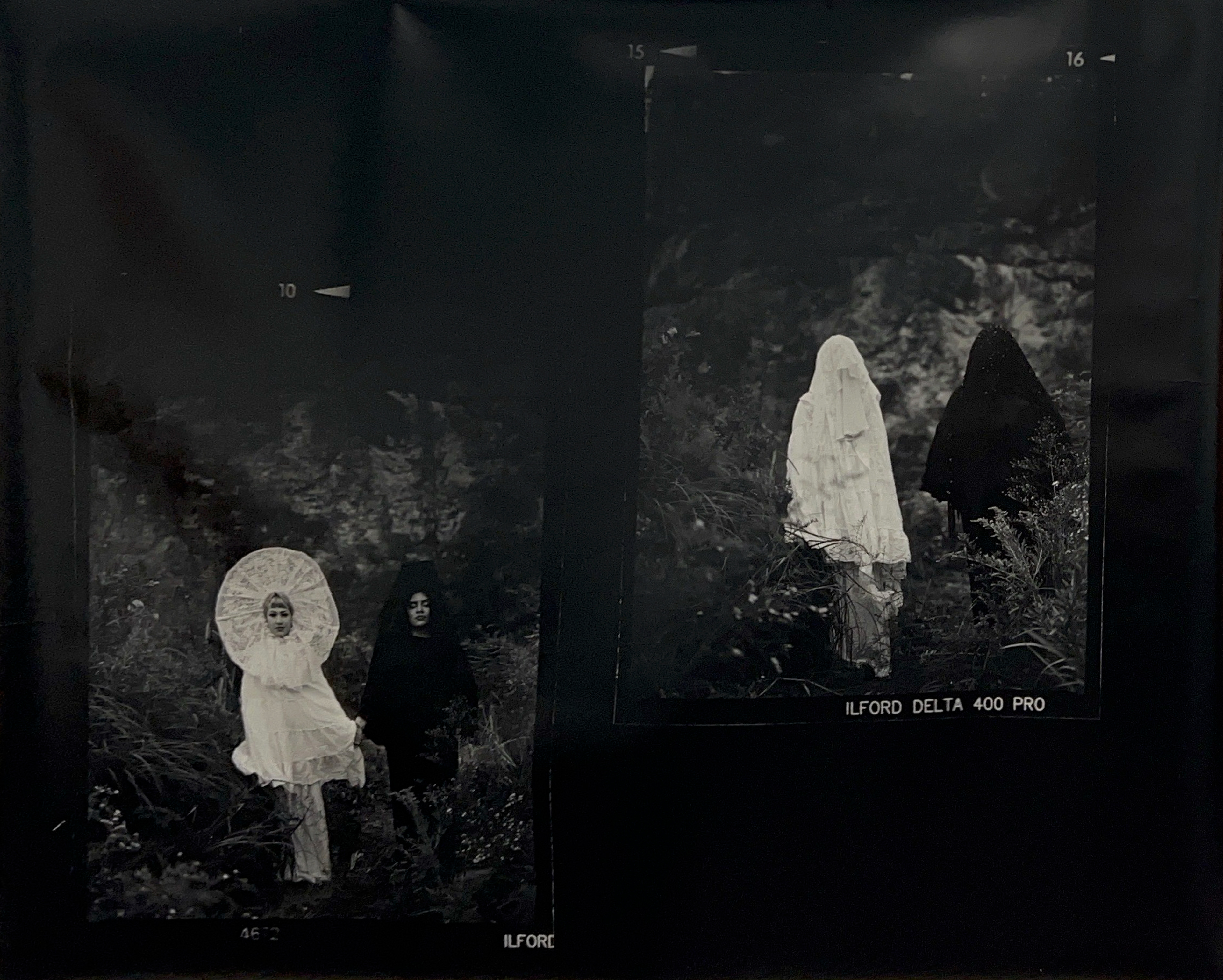 Black and white diptych photograph of two people standing amongst brush. One is wearing an all-white outfit and the other is wearing an all-black outfit. In the left photo the figures are looking at the camera. In the right photo, the figures' faces are covered by veils.