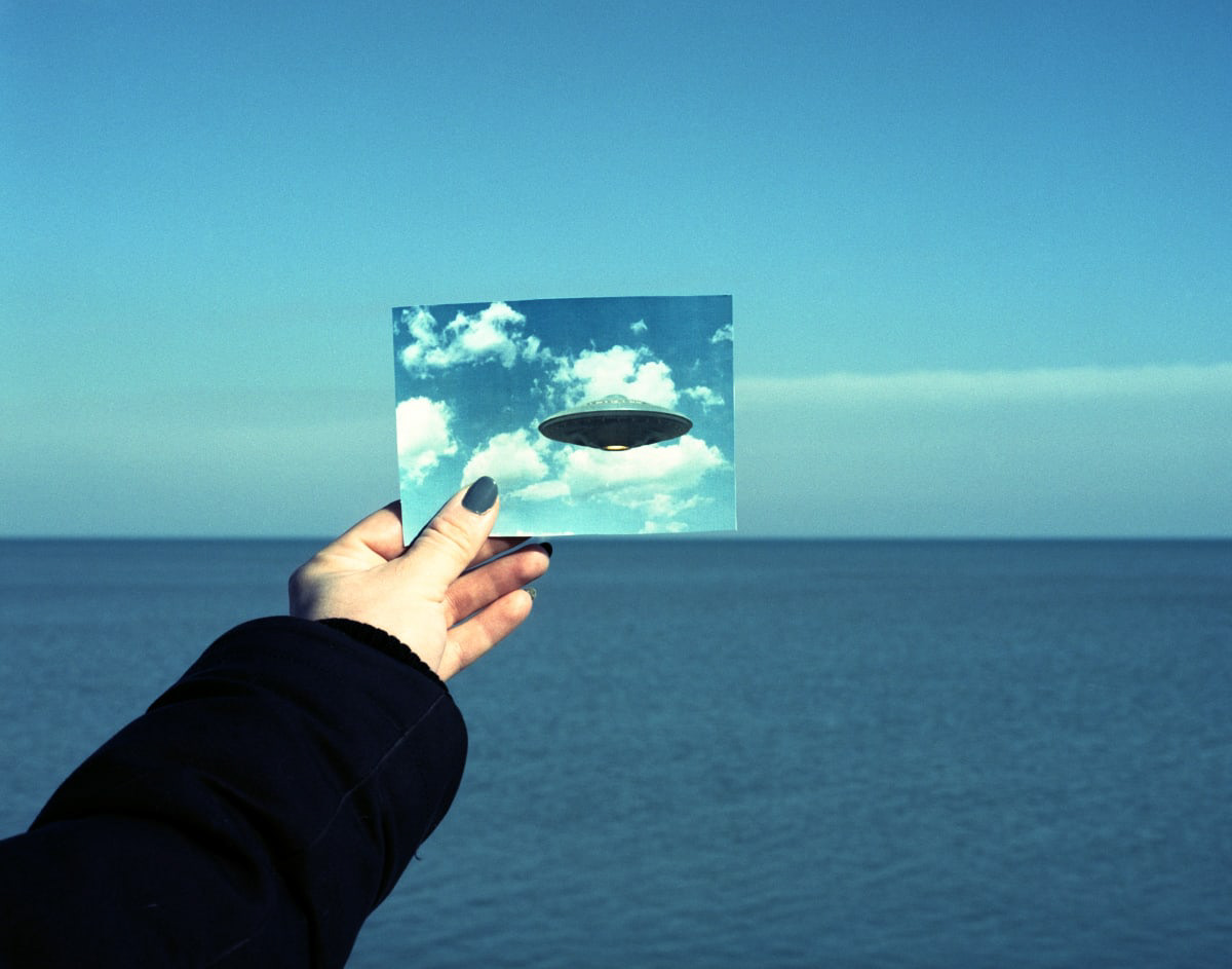Color photograph of an arm holding out a photo of a flying saucer in line with the horizon over a lake.