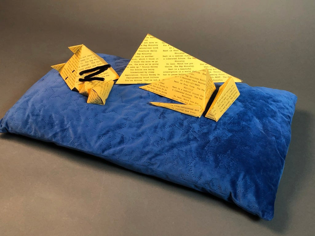 Yellow paper sculpture of a cat laying on a blue velvety pillow