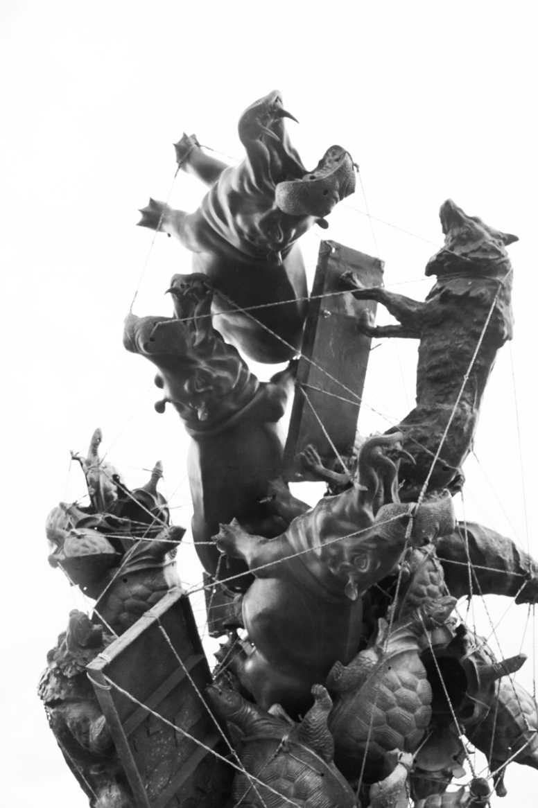 Black and white photo of a jumble of animal statues and planks held together by wire against a white background