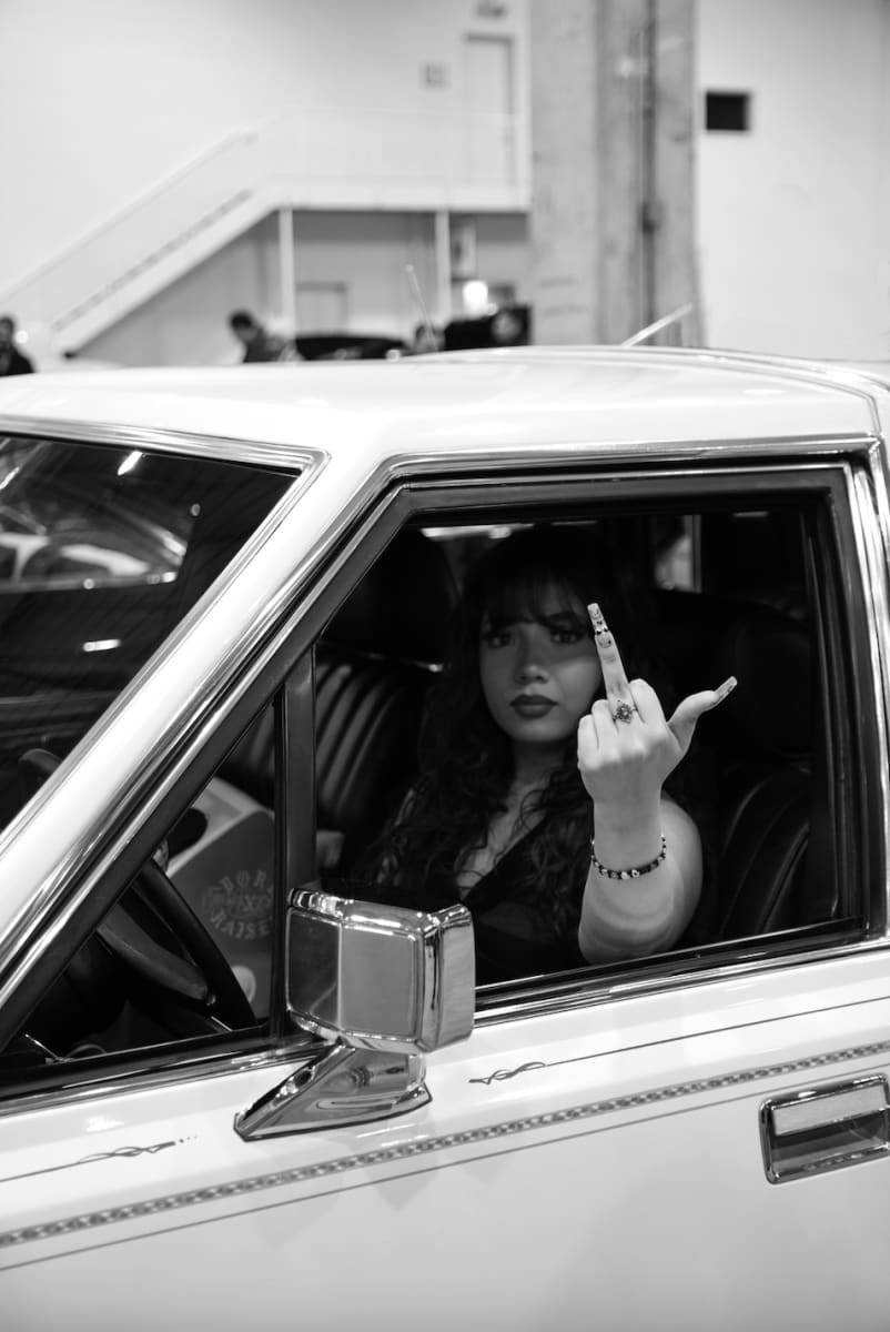 Black and white photo of a woman sitting in the drivers seat of a car with her middle finger up and pointed towards the camera