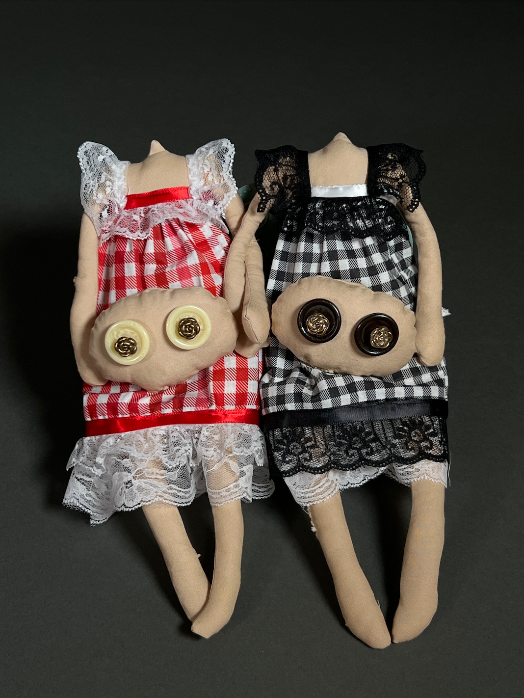 Two dolls hold their heads in their hands. The dolls have checkered dresses in red and black with white lace and the heads have button eyes.