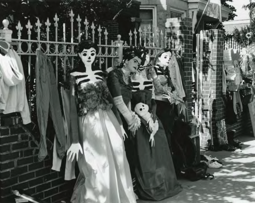 Black and white photo of skeletal and mannequin figures leaning against a wrought iron fence and brick wall
