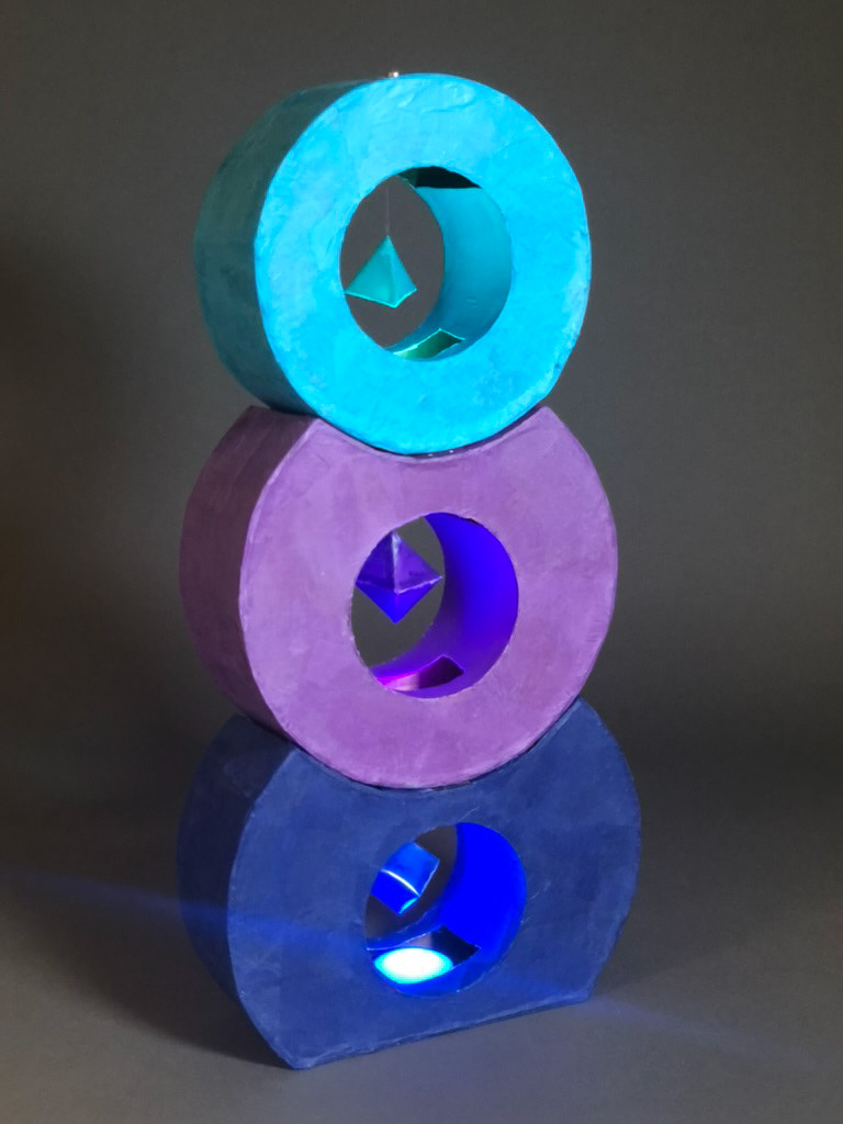 Sculpture of three stacked rings in dark blue, purple, and light blue colors. Each ring has a small shape suspended within it which is illuminated by a light below.