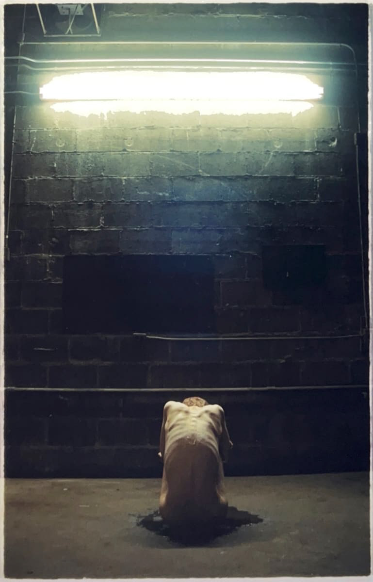 A photograph of a skinny man with bones visible beneath his skin sits hunched on the floor with his back to the camera and in front of a dark brick wall illuminated at the top by a long fluorescent light
