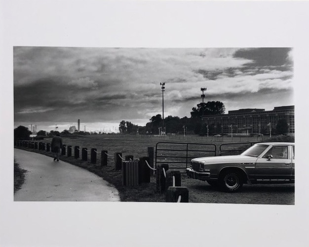 Black and white photograph of a parked car next to a field and sidewalk with a lone person walking