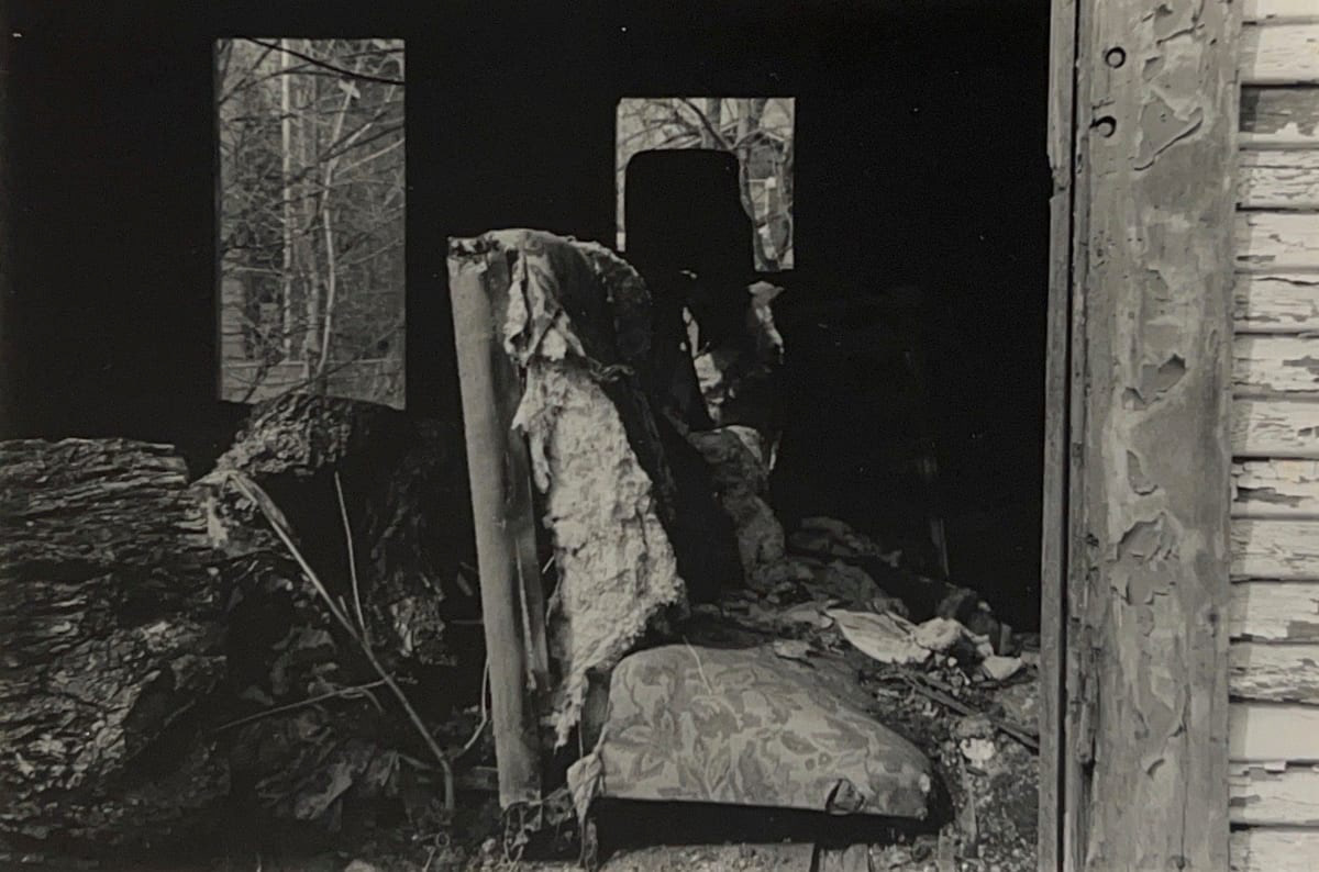 Black and white photograph of the interior of an abandoned house with a worn down chair in the foreground, surrounded by overgrowth