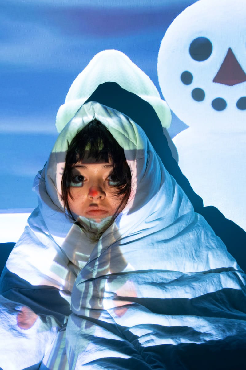 Color photograph of a woman wrapped in a comforter with an image of a cartoon person and snowman projected on top of her.