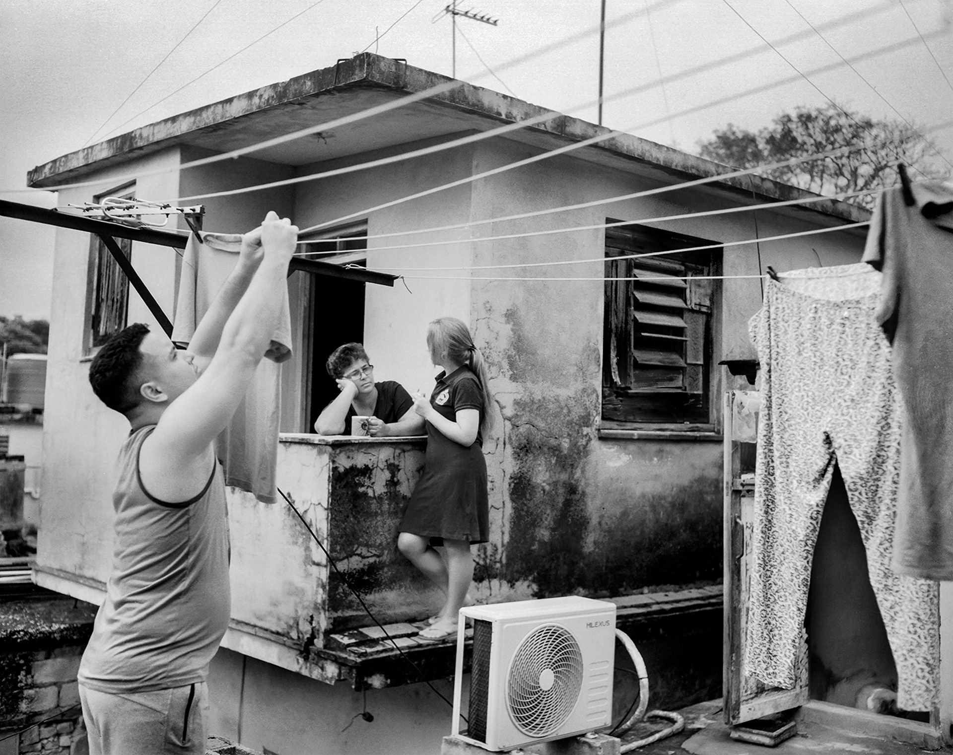 Black and white photograph of a man hanging laundry on a clothes lines and two women having a conversation in the background in front of a house.