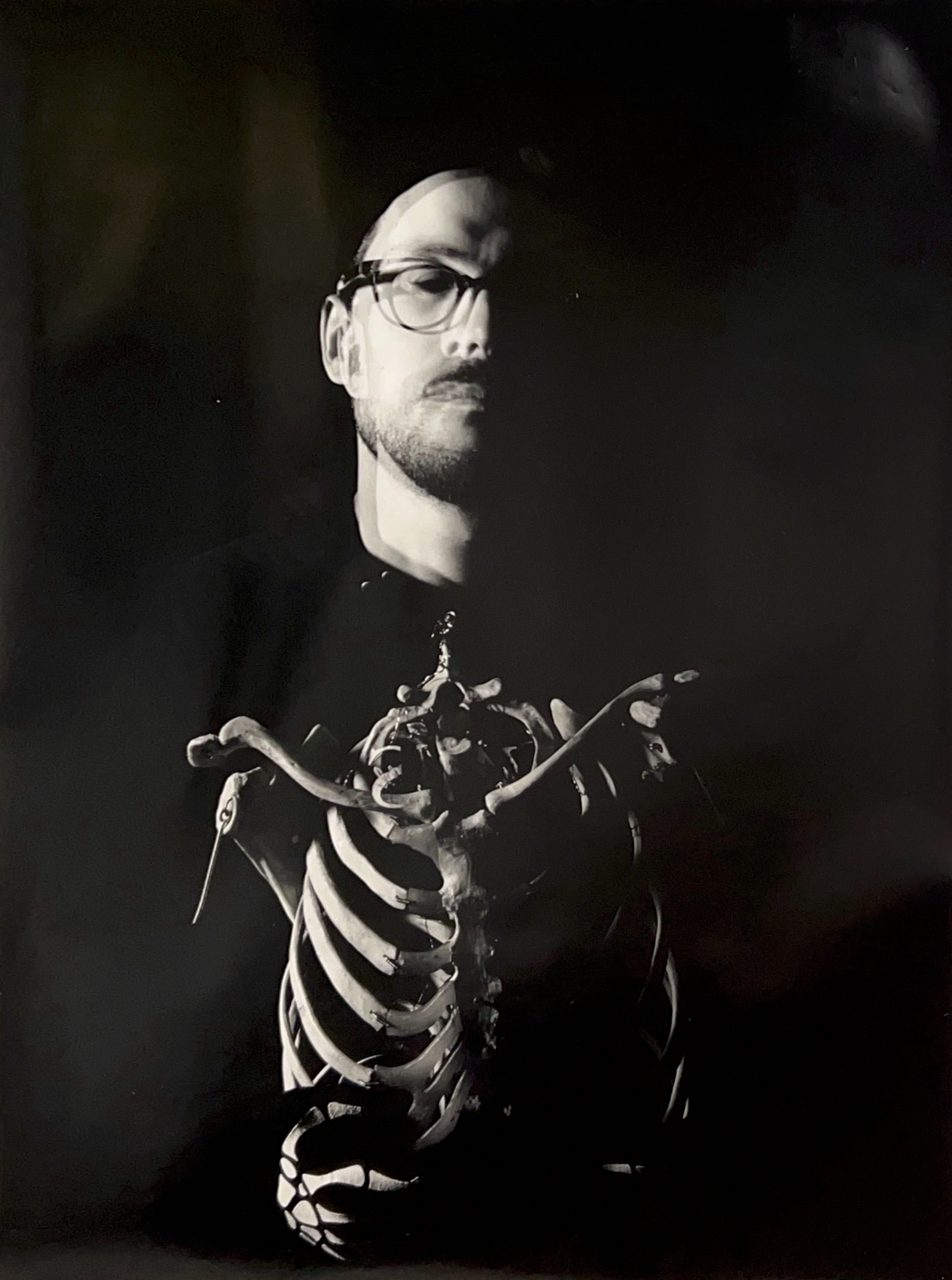 Black and white photograph of a man with a skeleton chest in front of him. The man is wearing glasses and half his face is shrouded in shadow.
