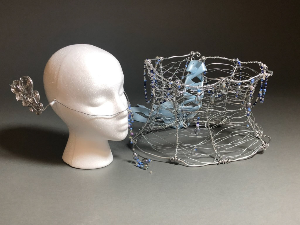 Wire corset with light blue lace and blue beading next to a foam head wearing wire face jewelry with blue beading.