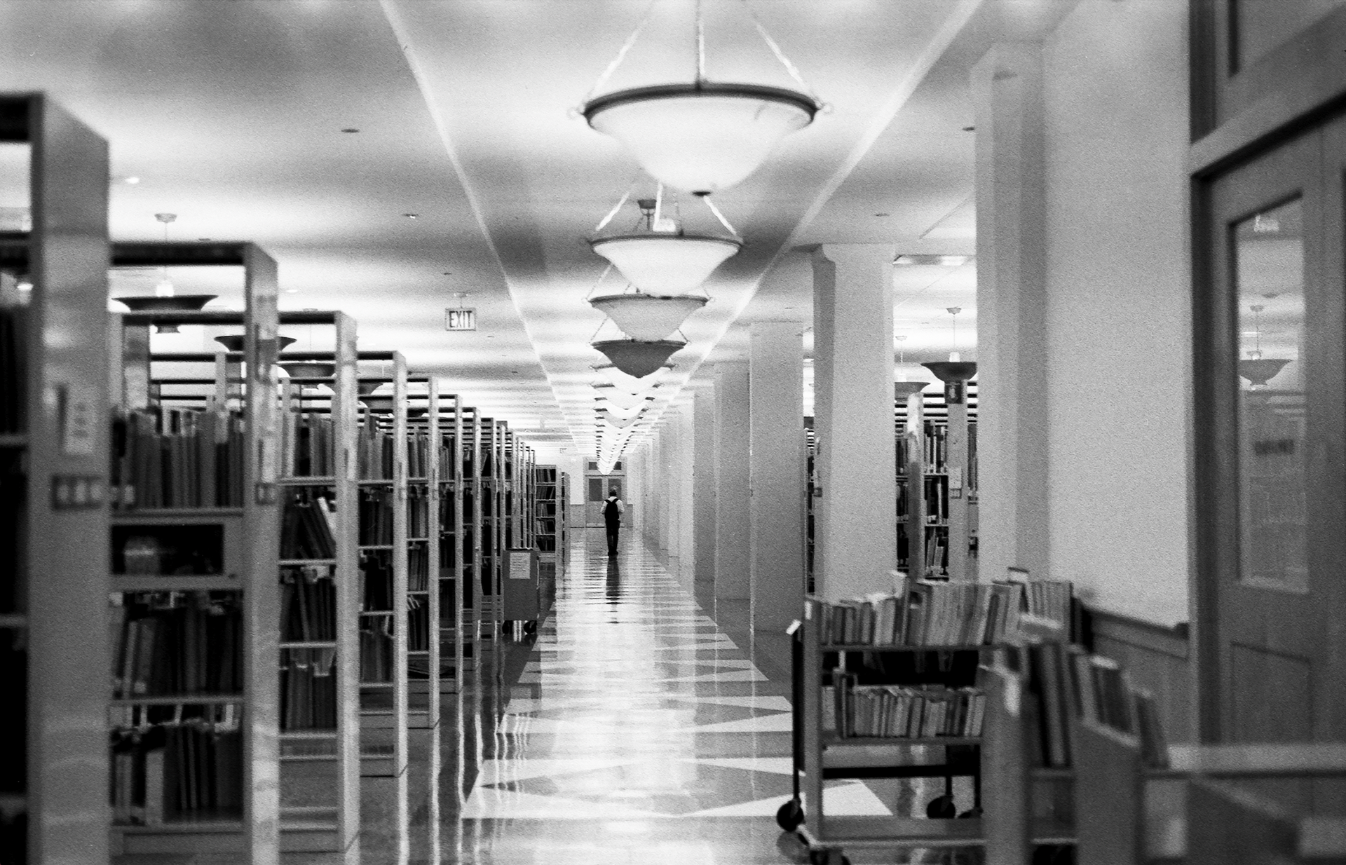 Black and white photograph of a library hallway with ceiling lamps, bookshelves, and a book cart. A lone figure walks toward the end of the hallway.