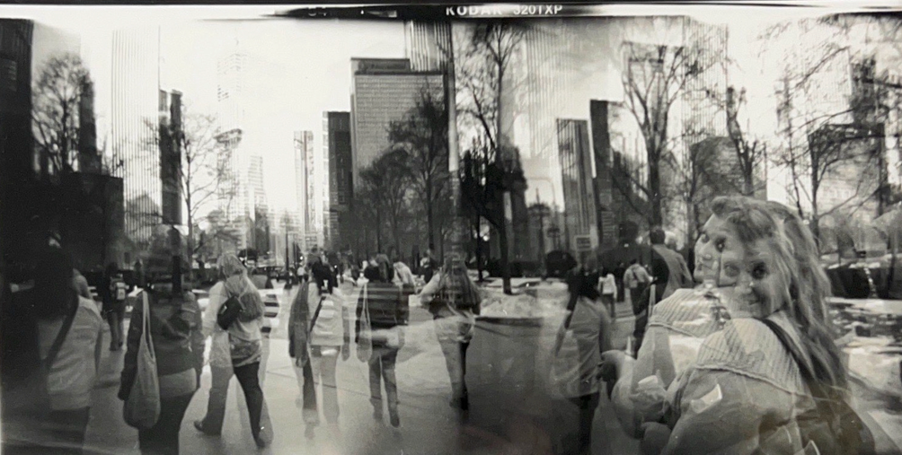 black and white photo of people walking in an urban setting. The photo has a long exposure, making the people look ghost-like.