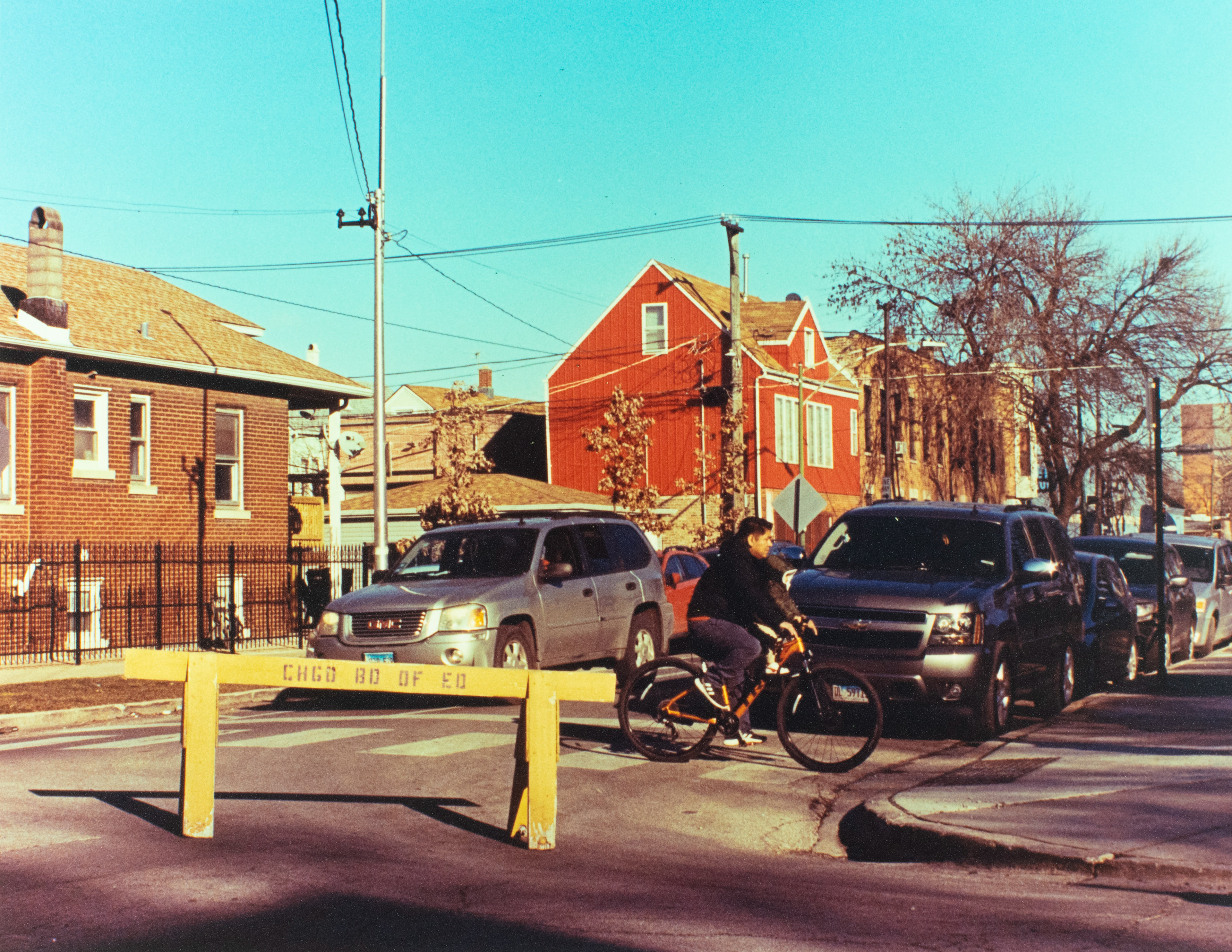 Photograph showing a bike-rider moving past parked cars and houses on a closed street.