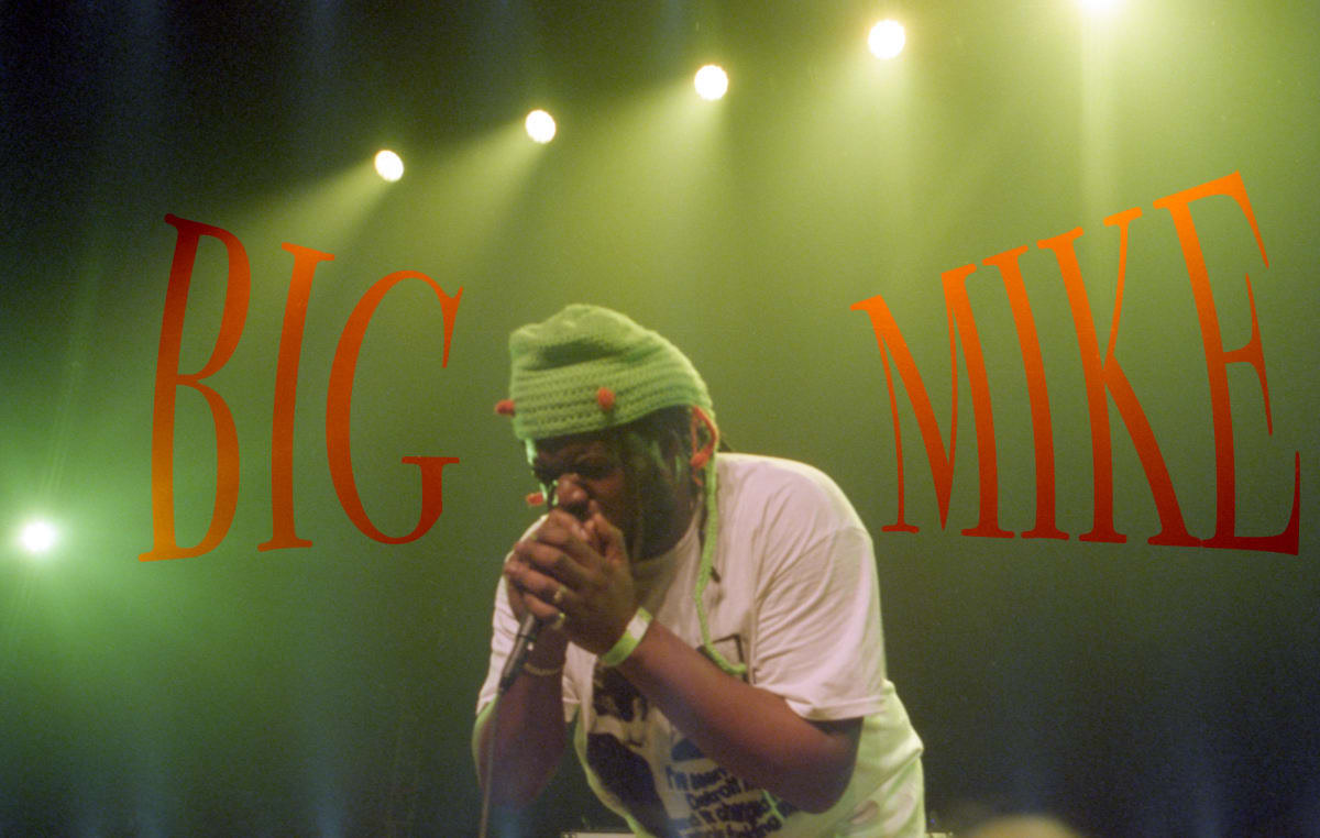 Photograph of a performer with a microphone under green lights with the words "Big Mike" written in orange text around him.