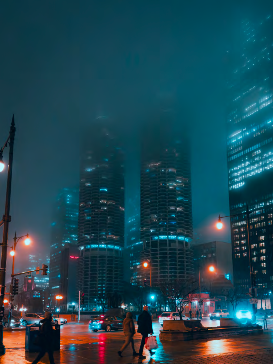 Street-view of city buildings partially shrouded by fog at night. People and cars move about at the bottom of the photo.