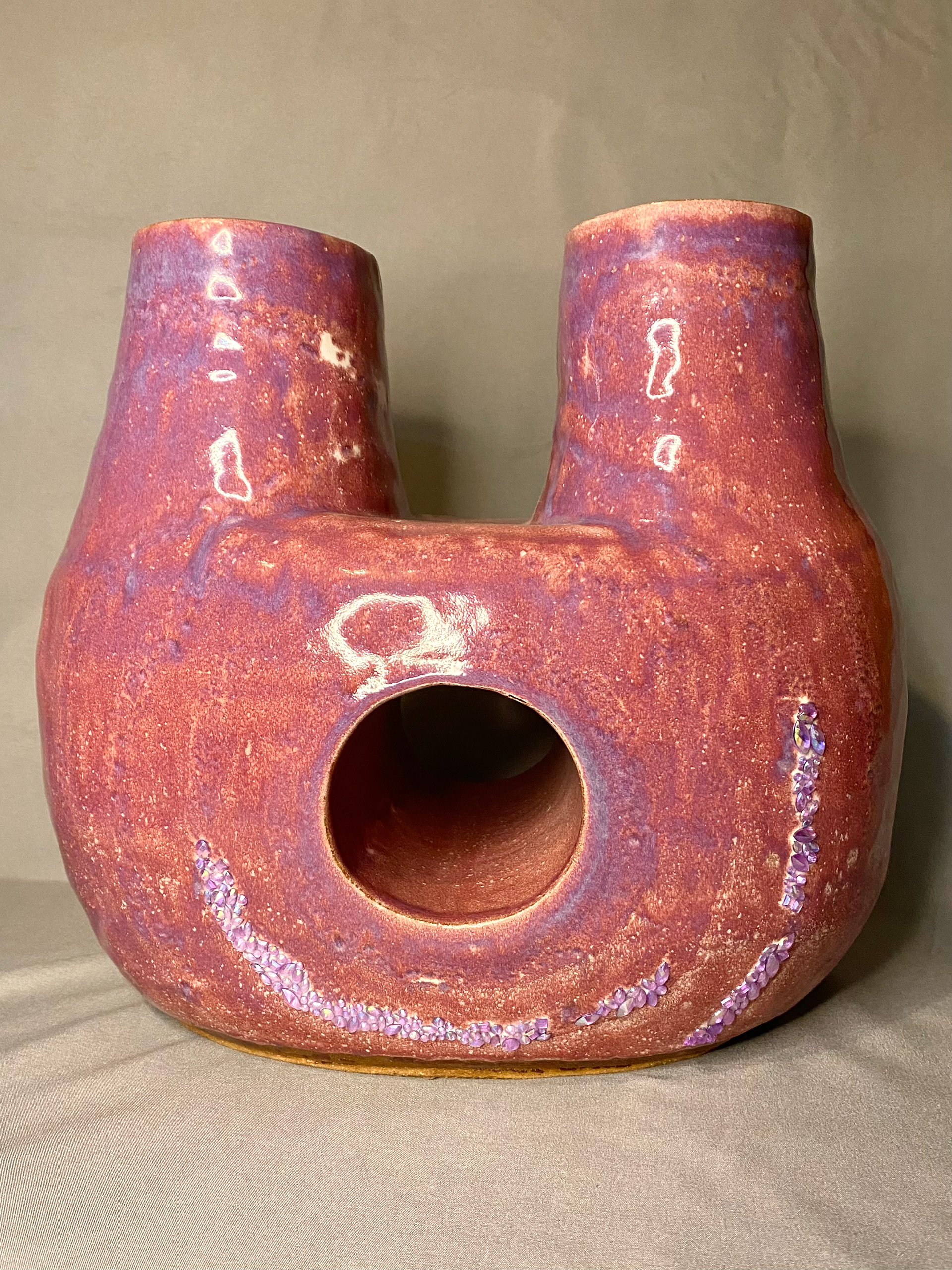 Pink ceramic vase with two heads and a hole through the middle. Sparkly gemstones are embedded in the vase along the bottom.