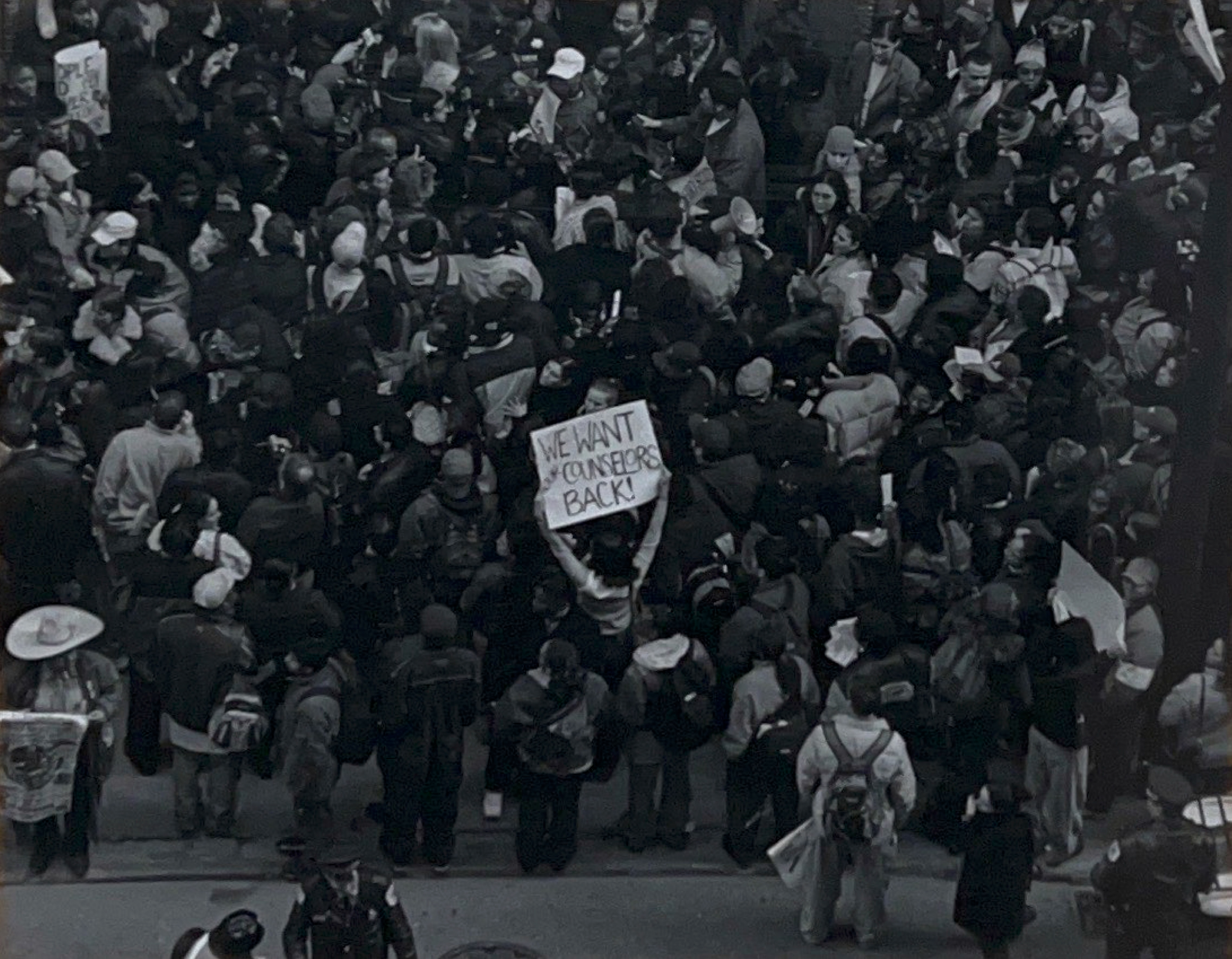 Black and white photograph of a crowd of people with one person holding a sign that reads "we want our counselors back"