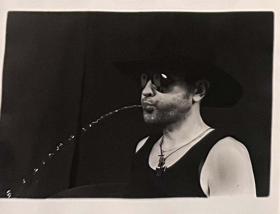 Black and white photograph of a man spitting a stream of water, wearing sunglasses, a rimmed hat, a tank top, and necklaces.