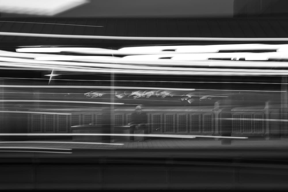 Long-exposure black and white photograph of a man sitting on a bench on a train platform.