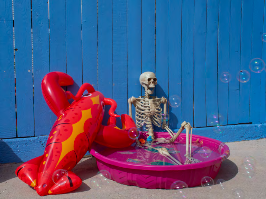A plastic skeleton and inflatable lobster sitting in a pink kiddie-pool, in front of a blue fence with bubbles floating in the foreground.