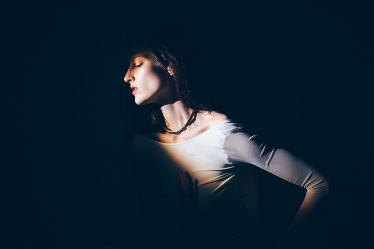 Portrait of a woman, partially shrouded in shadow and partially illuminated, looking off to the side and wearing a white, long-sleeved shirt and a black necklace.