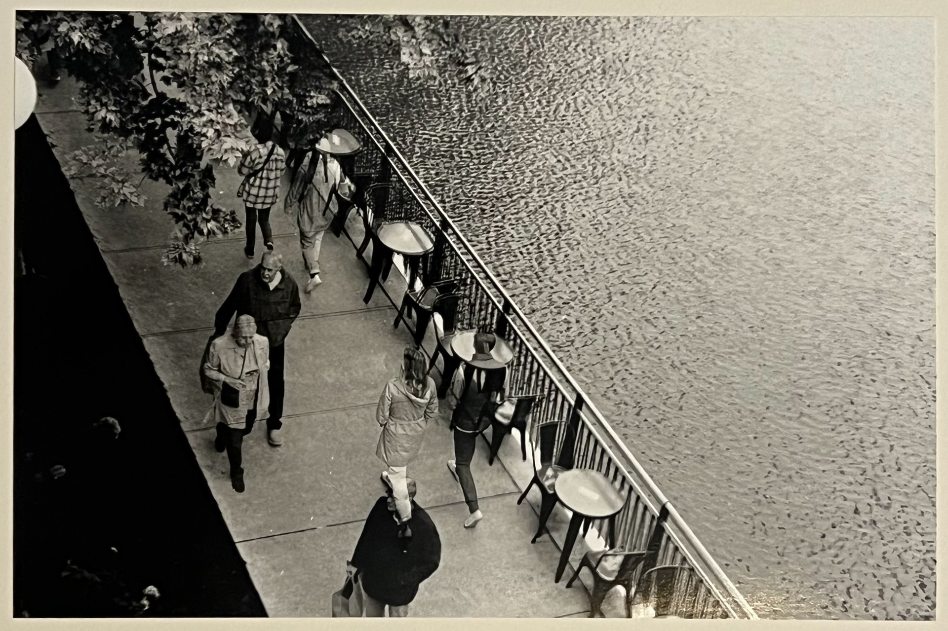 Black and white photo of pedestrians walking along a sidewalk overlooking water with tables and chairs lining the fence and tree branches hanging over the top lefthand corner of the image.