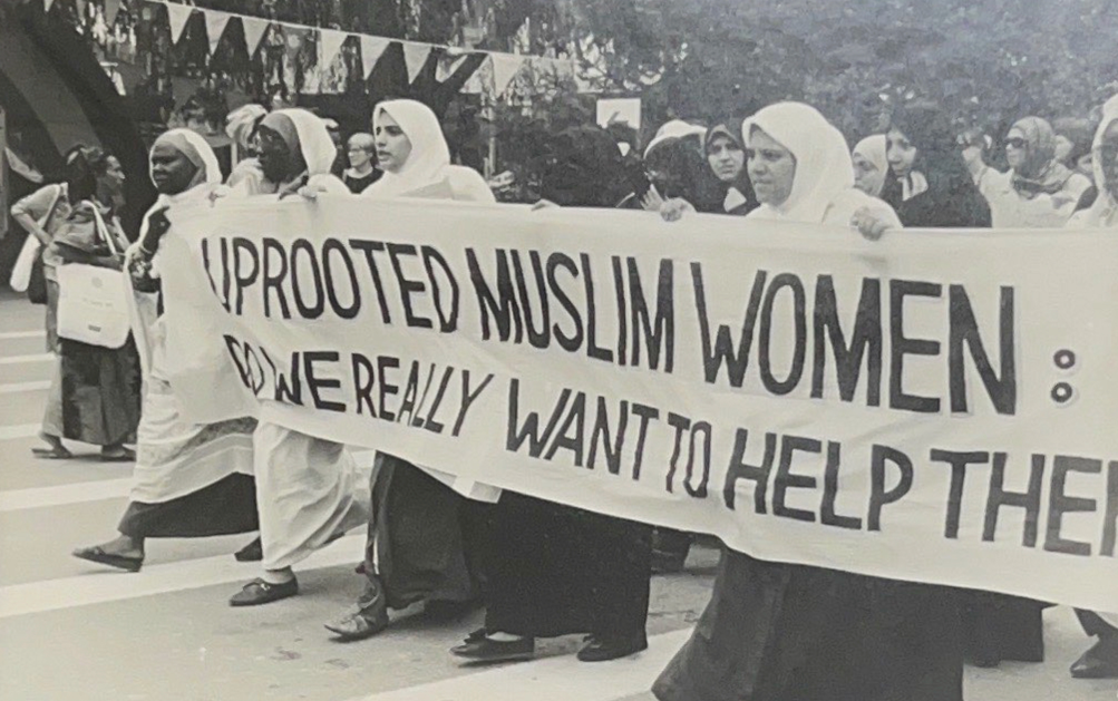 Black and white photo of a group of women marching and holding a large banner, partially cut off by the photo that reads "uprooted Muslim women: do we really want to help the"