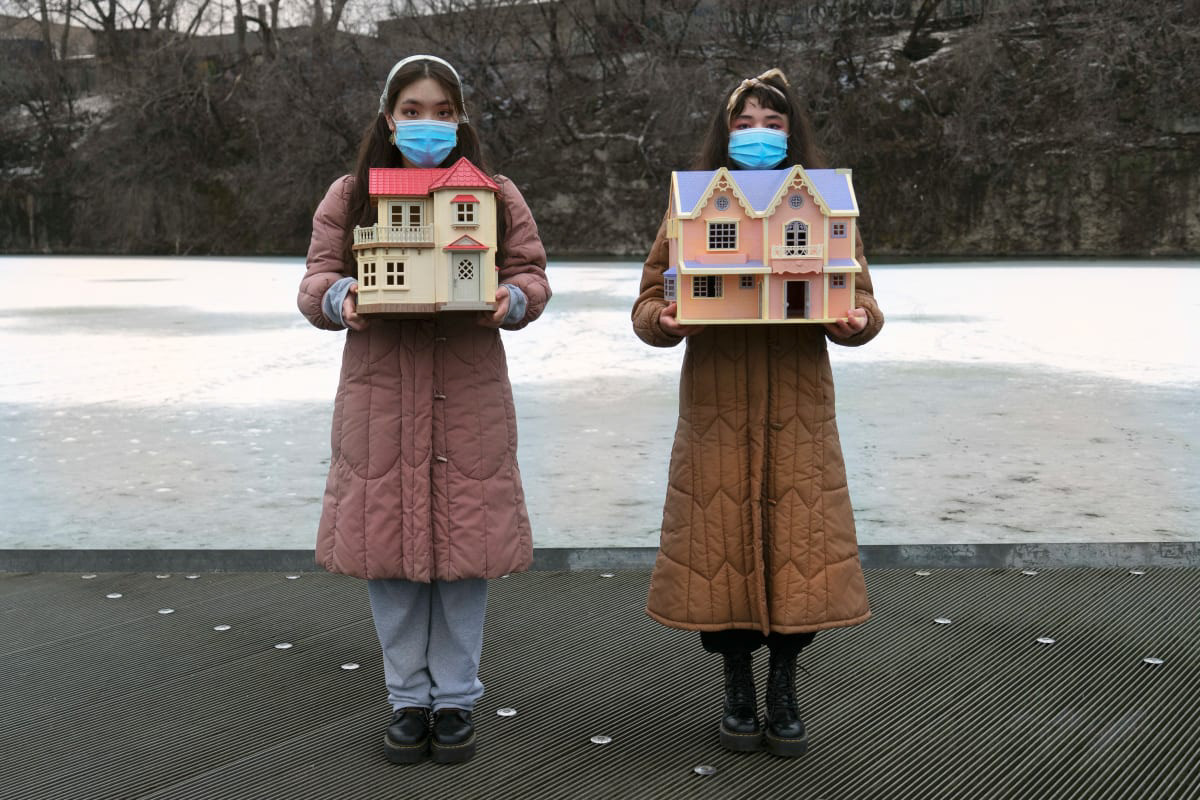 Color photograph of two women wearing long winter coats and blue surgical masks holding fanciful and detailed doll houses
