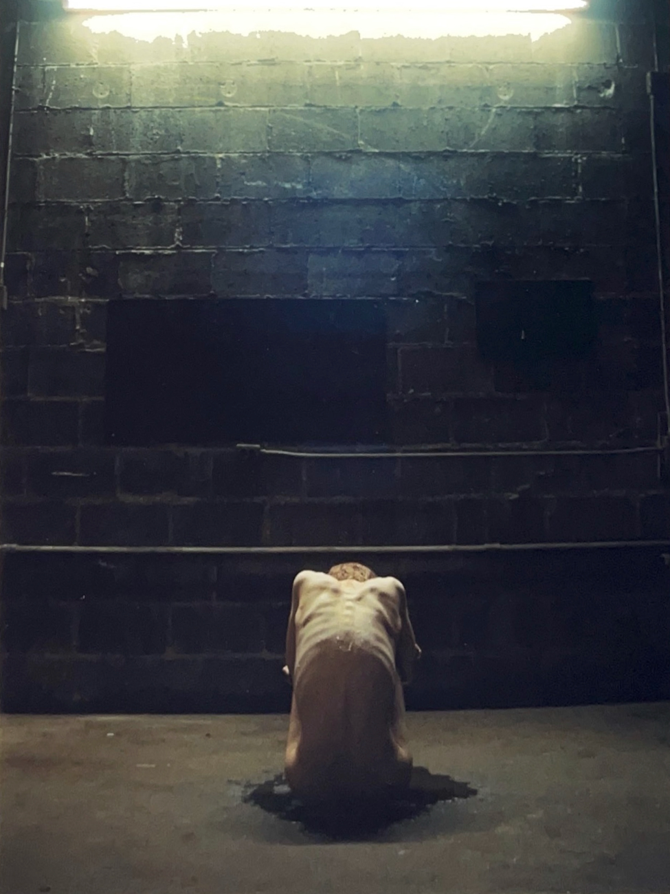 A photograph of a skinny man with bones visible beneath his skin sits hunched on the floor with his back to the camera and in front of a dark brick wall illuminated at the top by a long fluorescent light