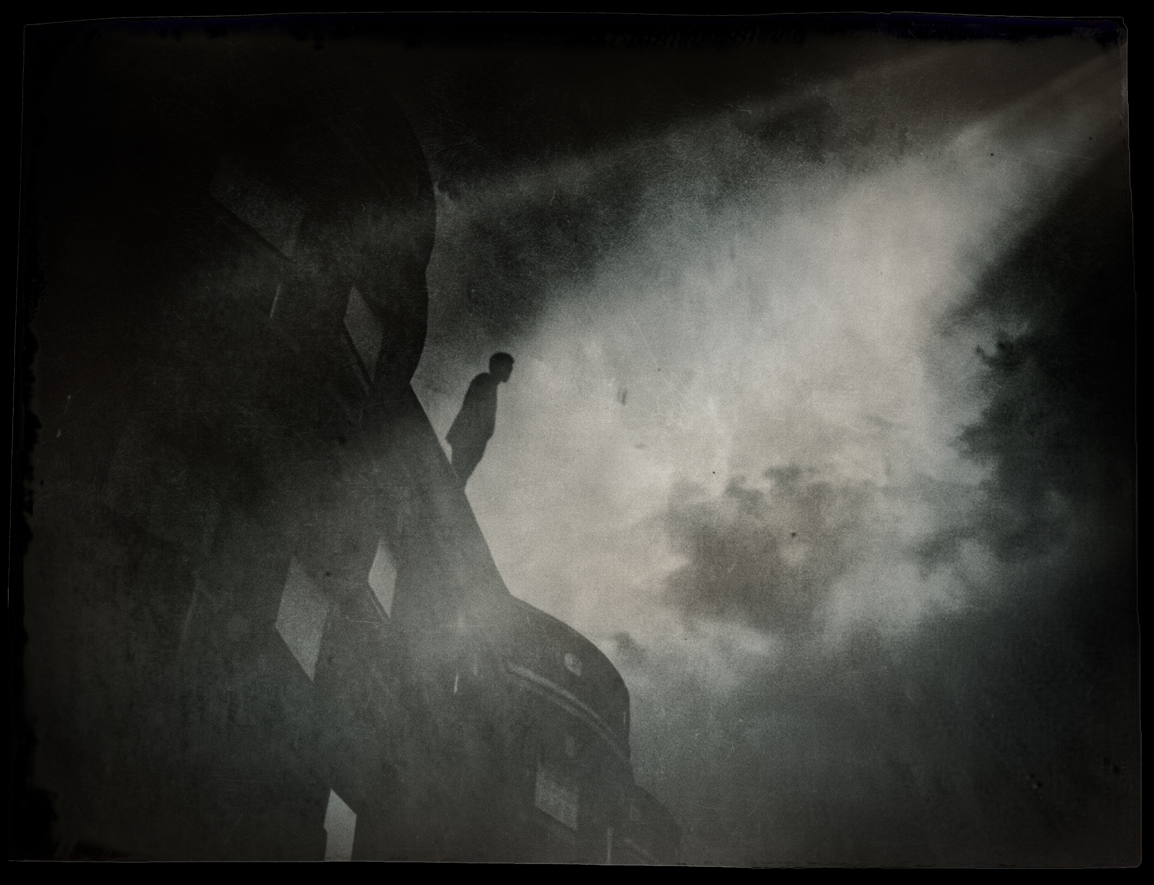 A man standing at the edge of a dark building's roof, in a spotlight of light from the sky