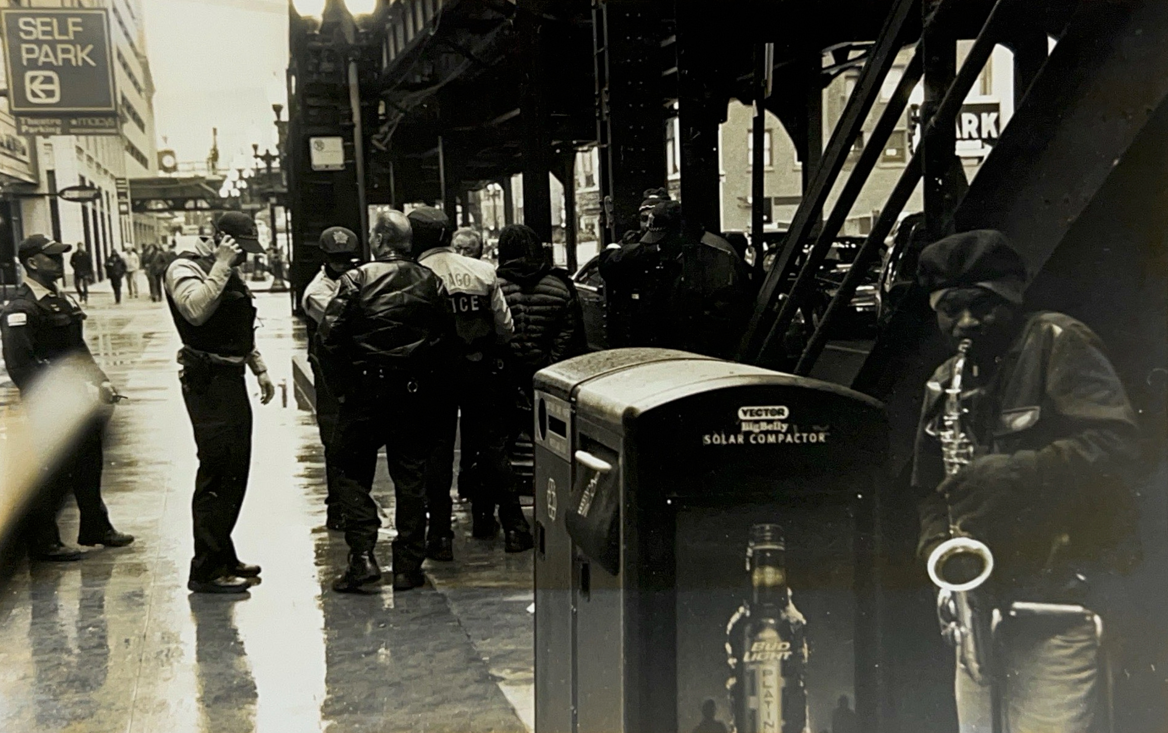 Black and white photograph of a Chicago street with L train tracks, a group of police officers congregating in the background and a saxophone player in the foreground