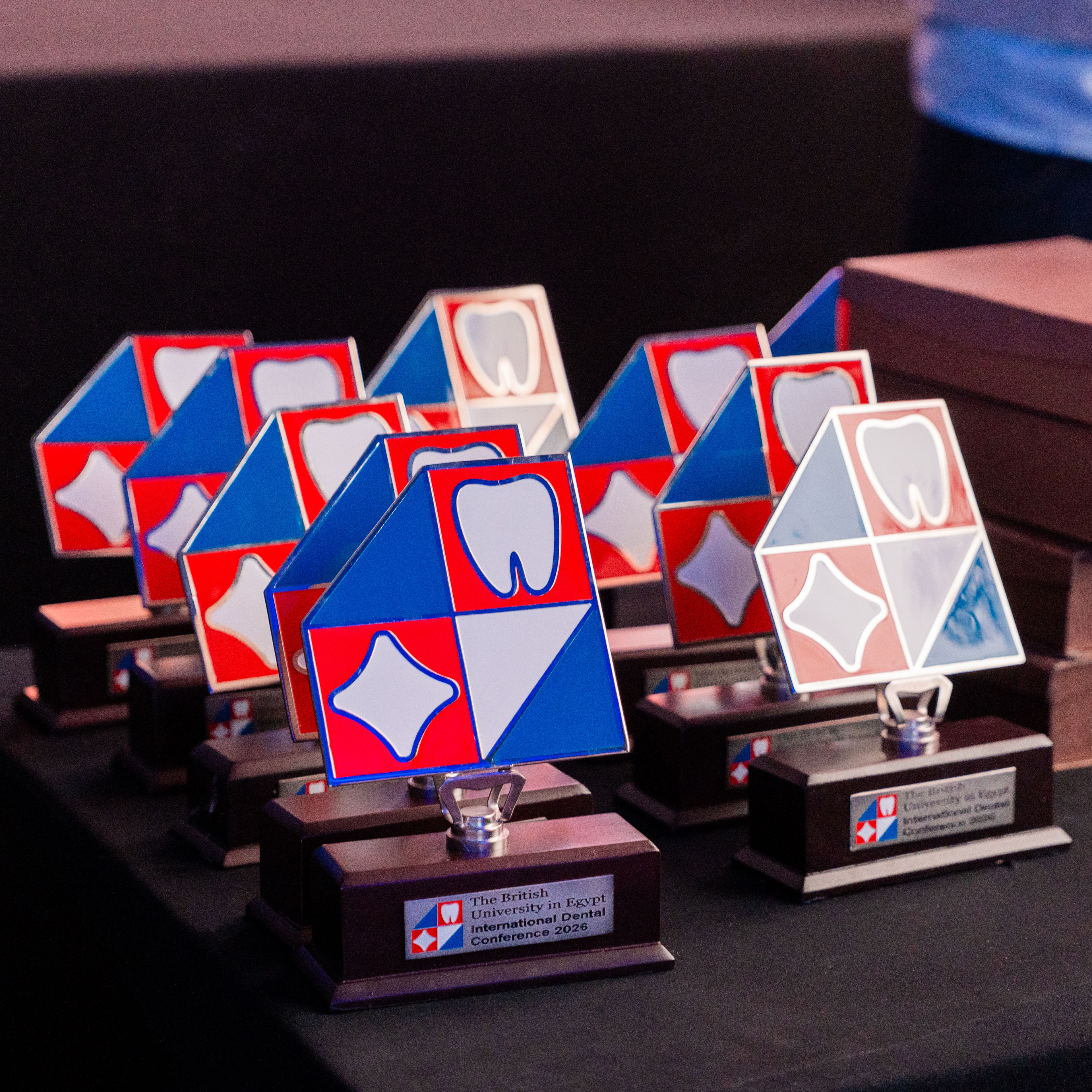 A row of custom-designed, modern geometric trophies by TheKconcept for The British University in Egypt International Dental Conference 2026, featuring a stylized tooth logo and a bold red, blue, and white color palette on premium dark wooden bases.